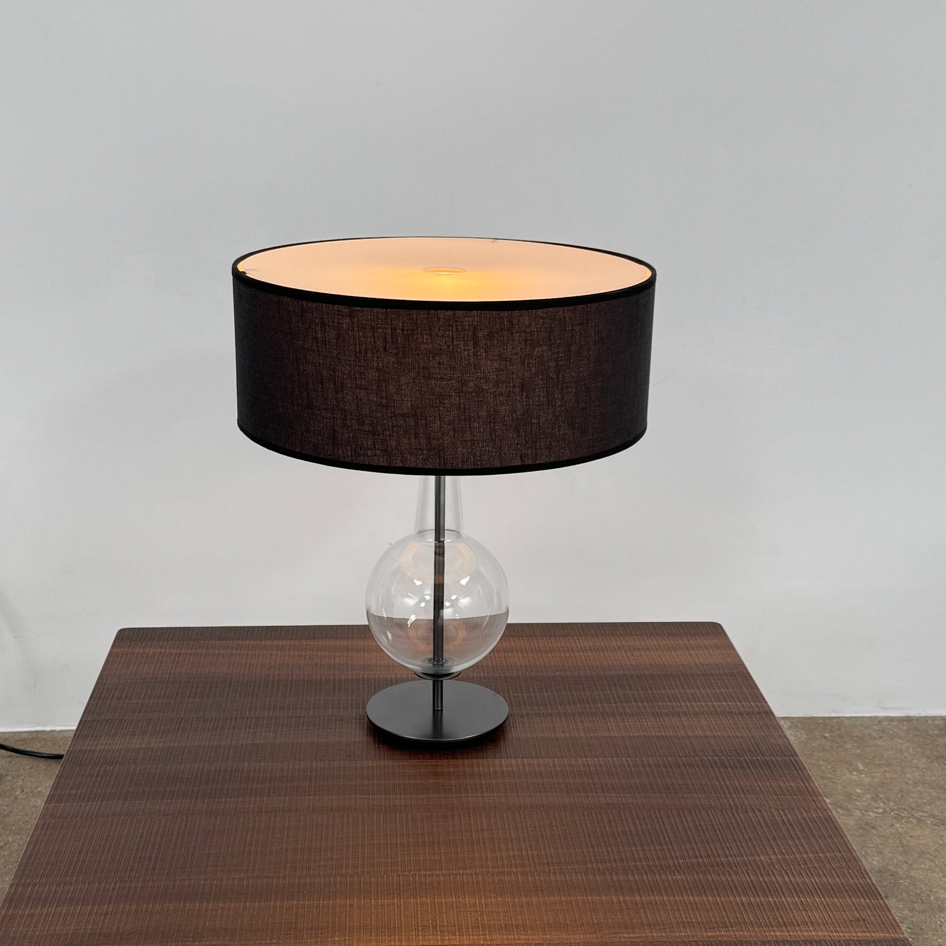 The Penta Classic Desir Table Lamp by Penta features a hand-blown glass base and a large cylindrical black lampshade, showcased against a plain white background.