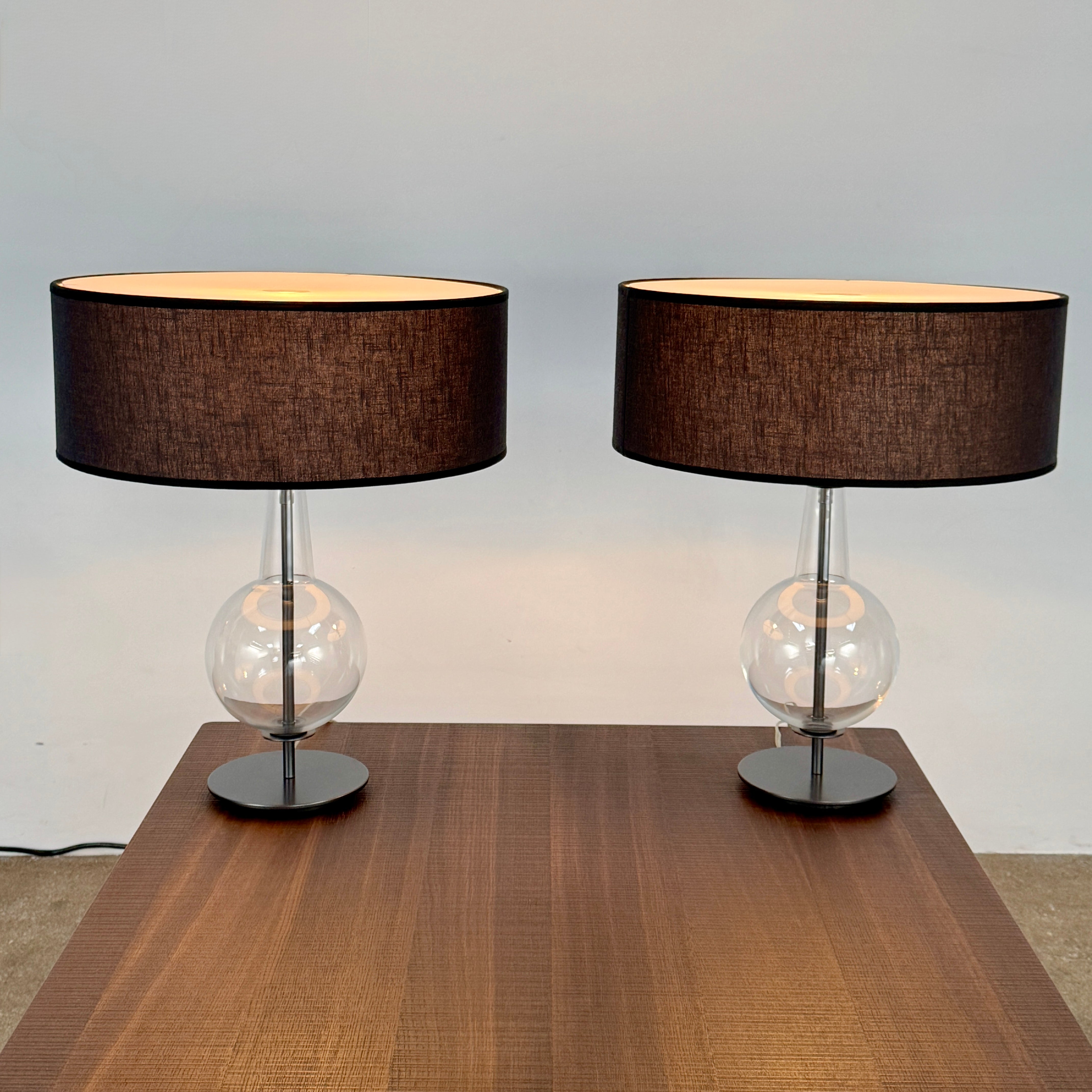 The Penta Classic Desir Table Lamp by Penta features a hand-blown glass base and a large cylindrical black lampshade, showcased against a plain white background.