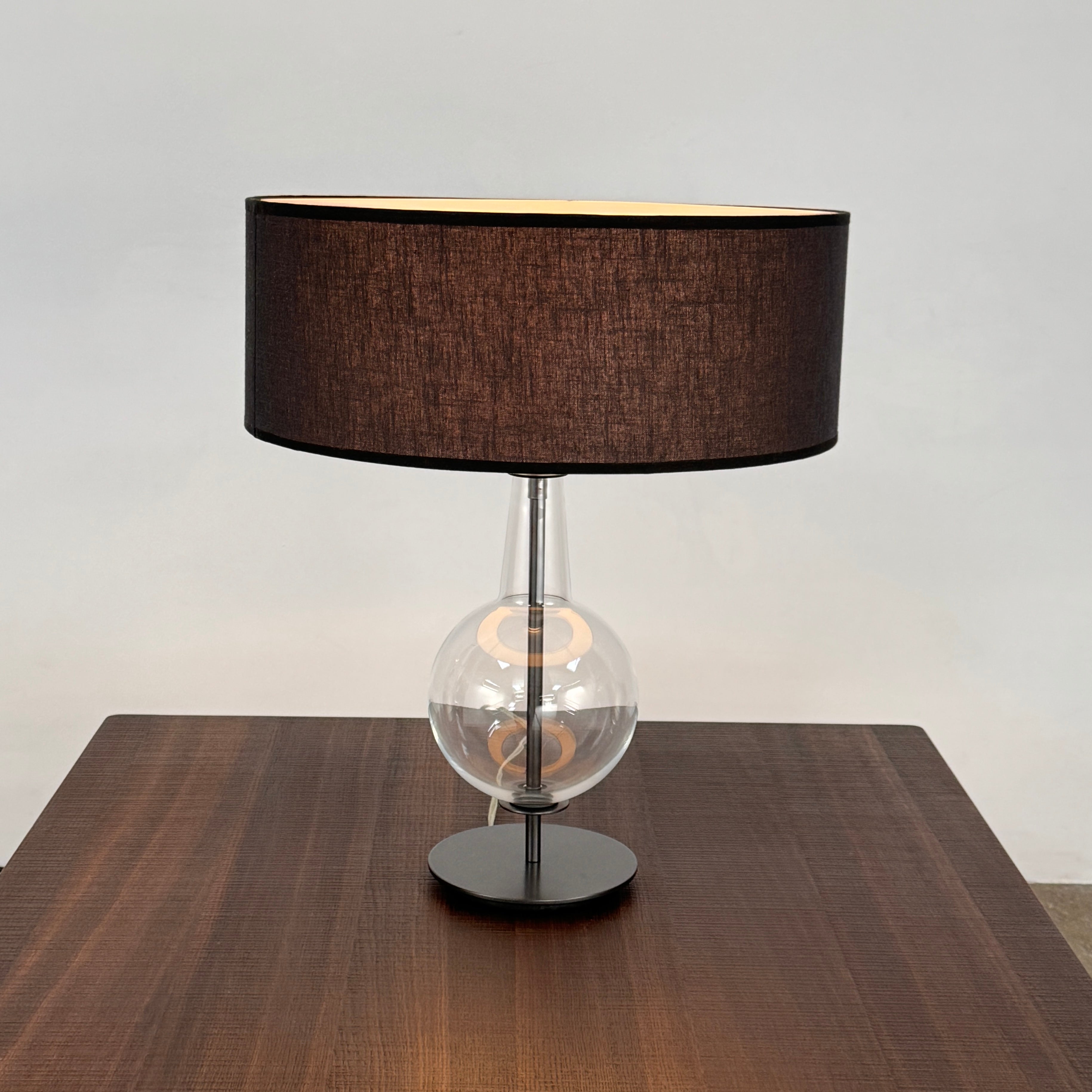 The Penta Classic Desir Table Lamp by Penta features a hand-blown glass base and a large cylindrical black lampshade, showcased against a plain white background.