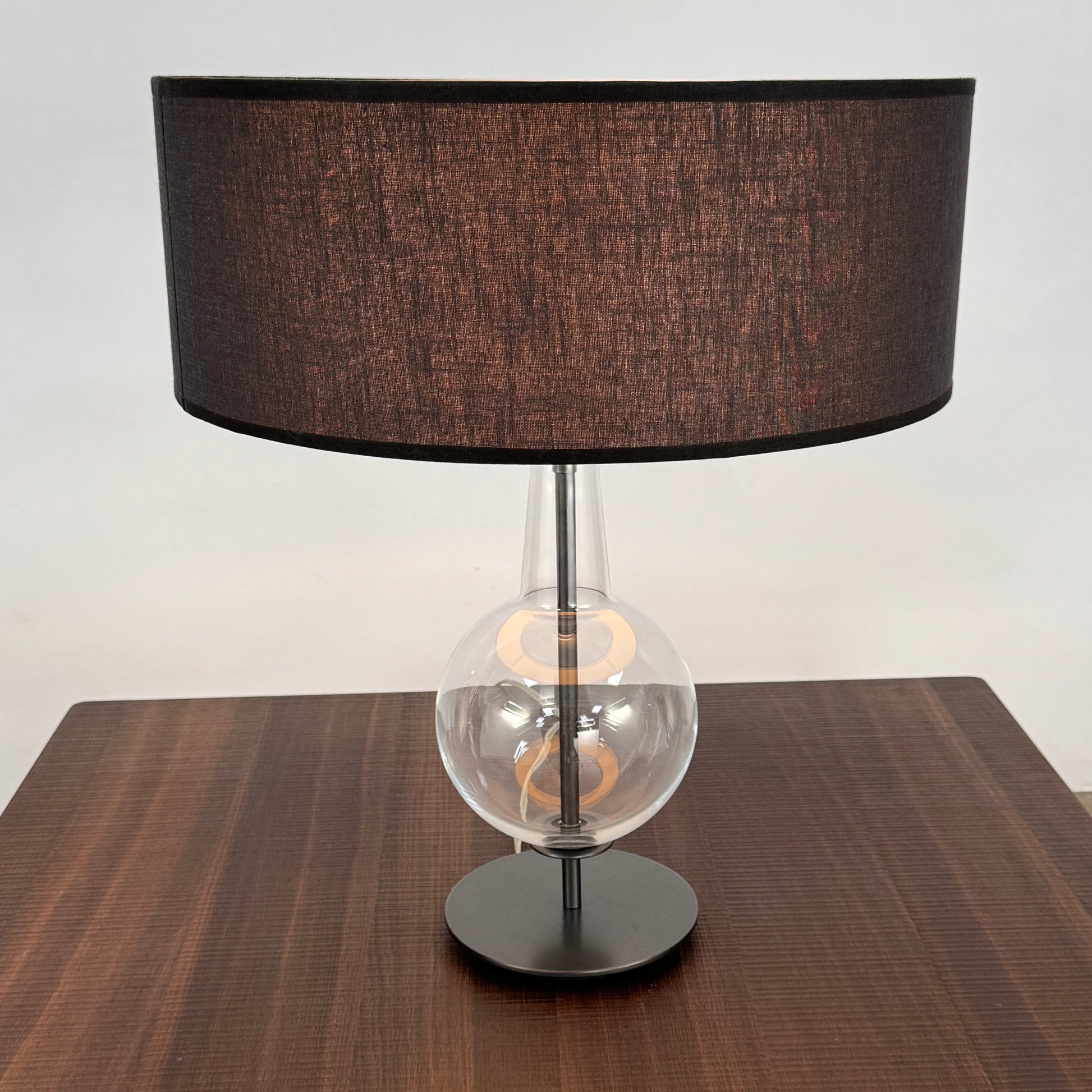 The Penta Classic Desir Table Lamp by Penta features a hand-blown glass base and a large cylindrical black lampshade, showcased against a plain white background.