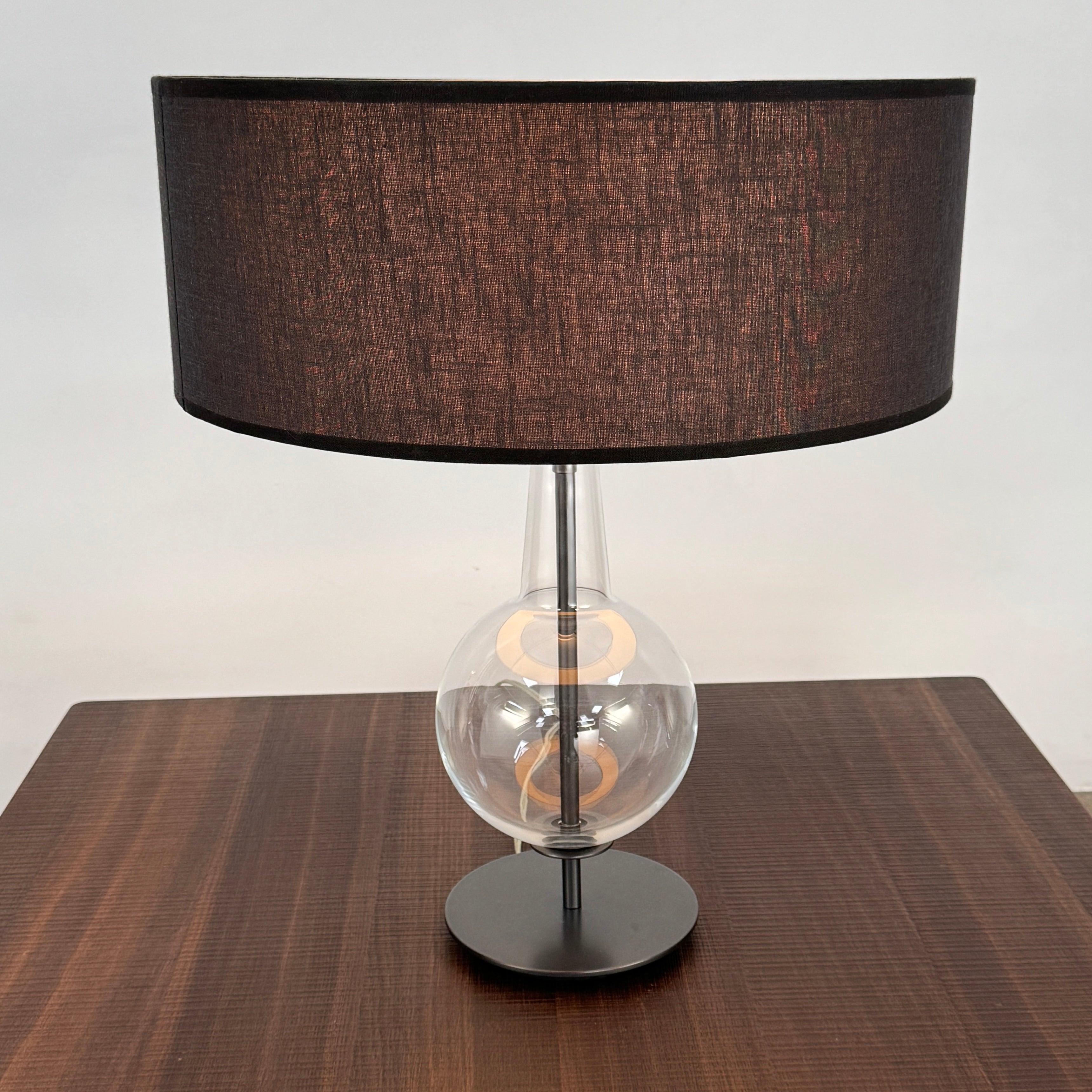 The Penta Classic Desir Table Lamp by Penta features a hand-blown glass base and a large cylindrical black lampshade, showcased against a plain white background.