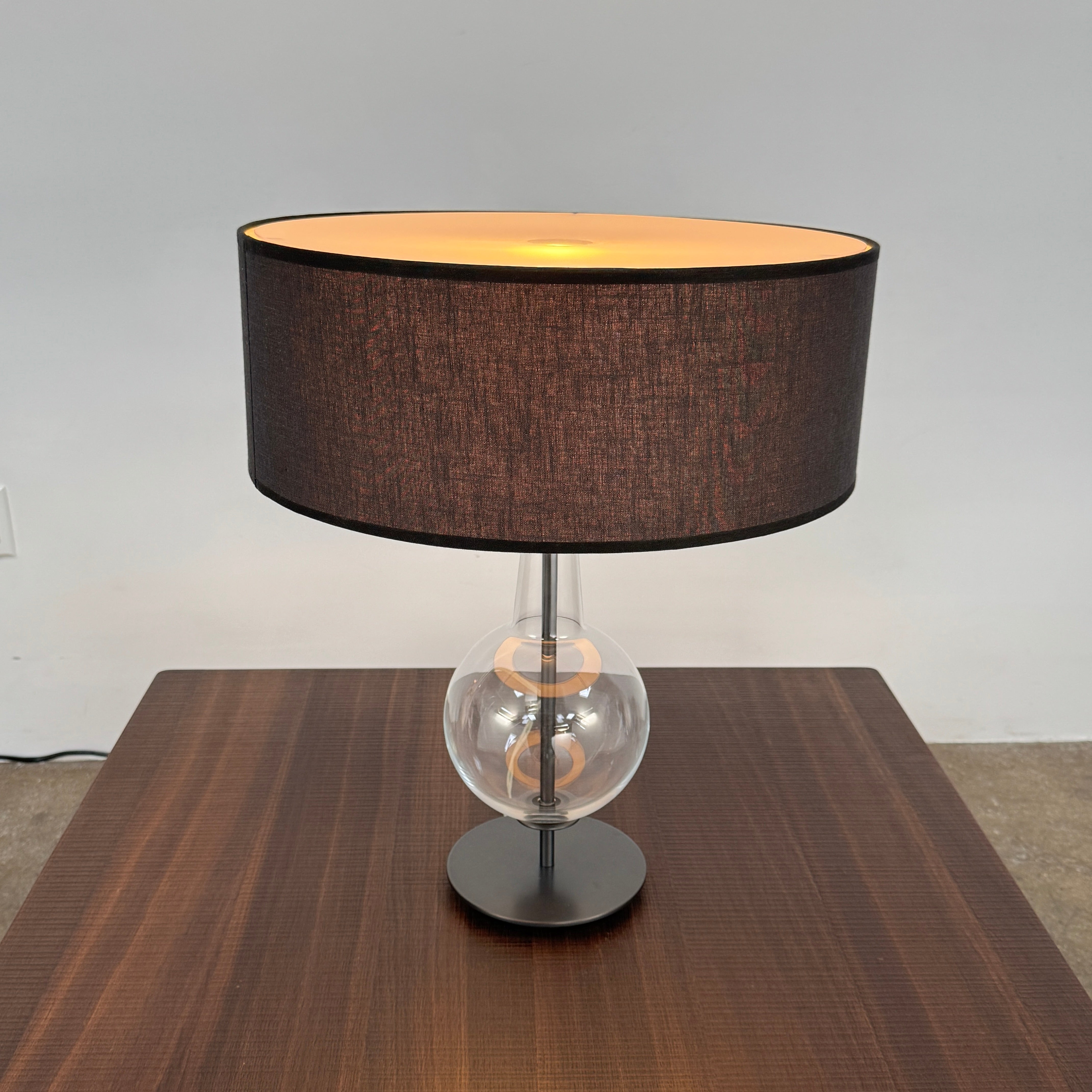 The Penta Classic Desir Table Lamp by Penta features a hand-blown glass base and a large cylindrical black lampshade, showcased against a plain white background.