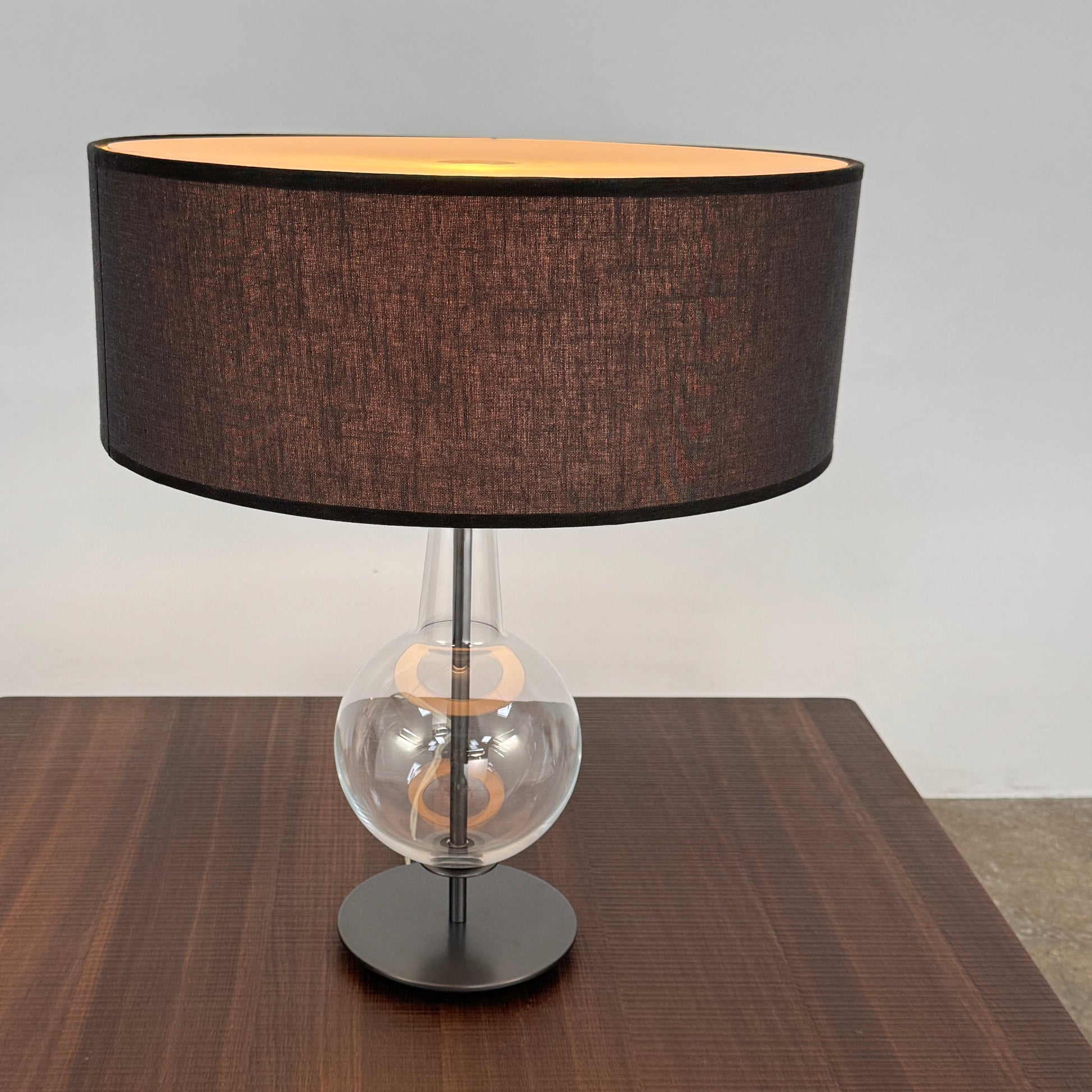 The Penta Classic Desir Table Lamp by Penta features a hand-blown glass base and a large cylindrical black lampshade, showcased against a plain white background.