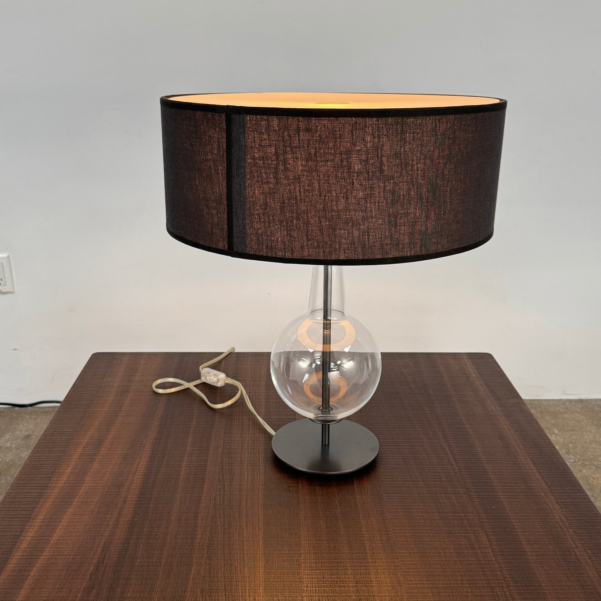 The Penta Classic Desir Table Lamp by Penta features a hand-blown glass base and a large cylindrical black lampshade, showcased against a plain white background.