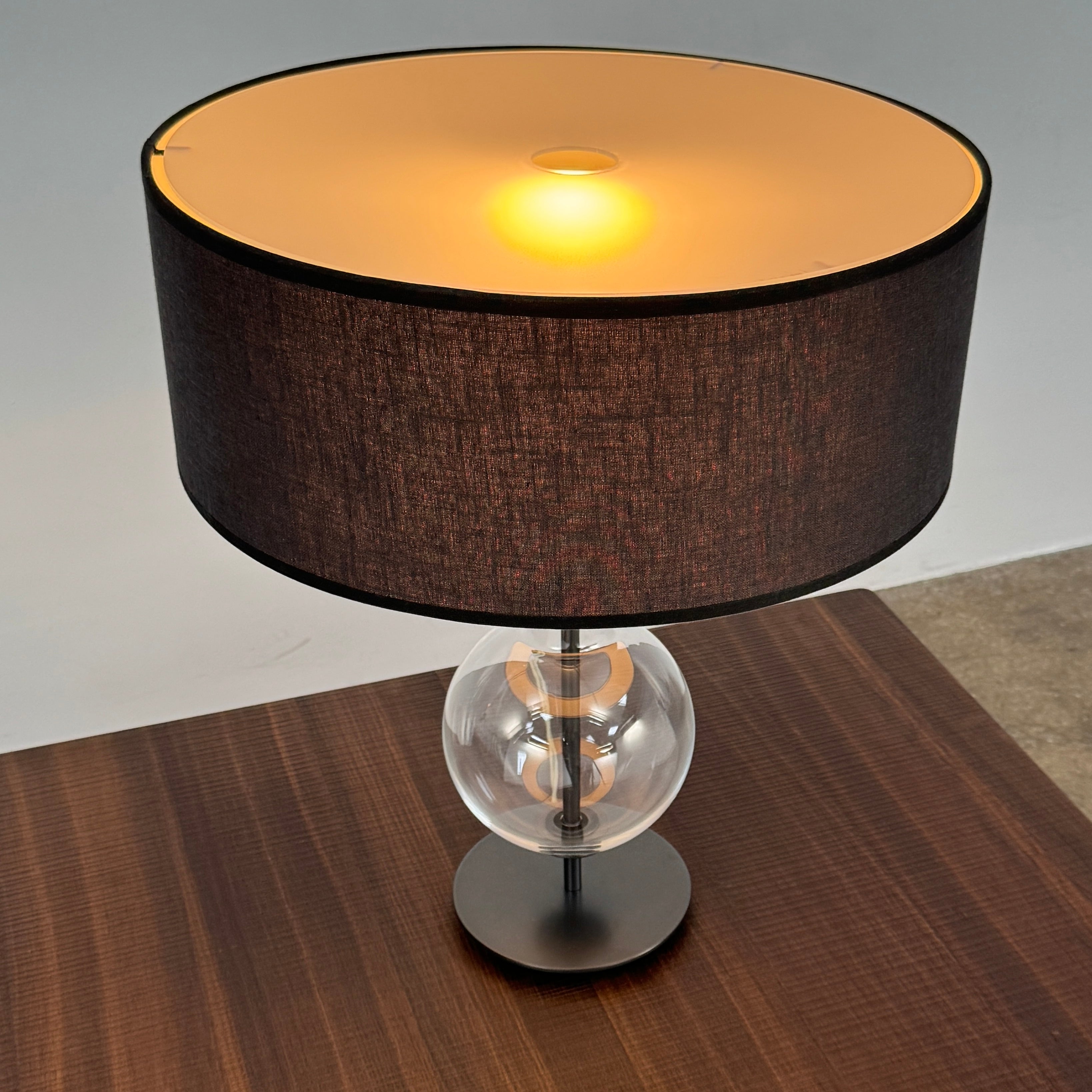 The Penta Classic Desir Table Lamp by Penta features a hand-blown glass base and a large cylindrical black lampshade, showcased against a plain white background.