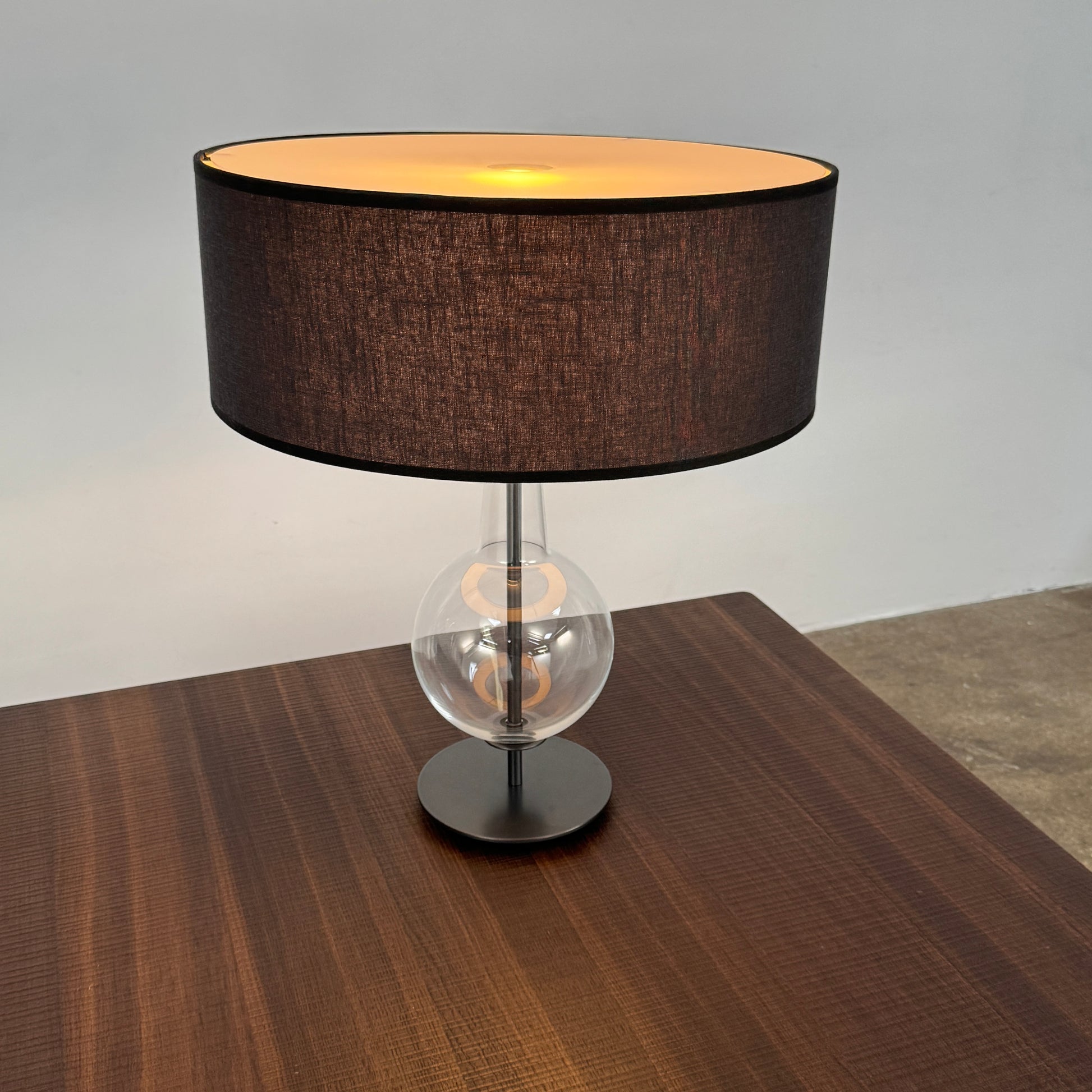 The Penta Classic Desir Table Lamp by Penta features a hand-blown glass base and a large cylindrical black lampshade, showcased against a plain white background.