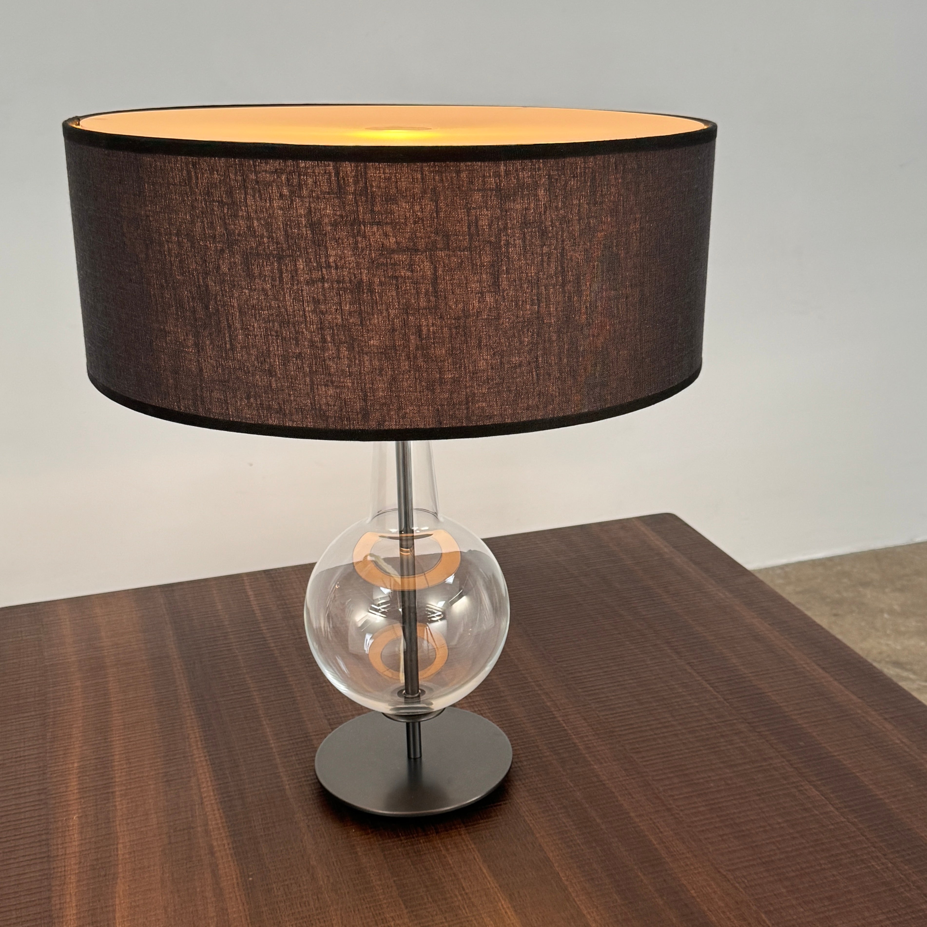 The Penta Classic Desir Table Lamp by Penta features a hand-blown glass base and a large cylindrical black lampshade, showcased against a plain white background.