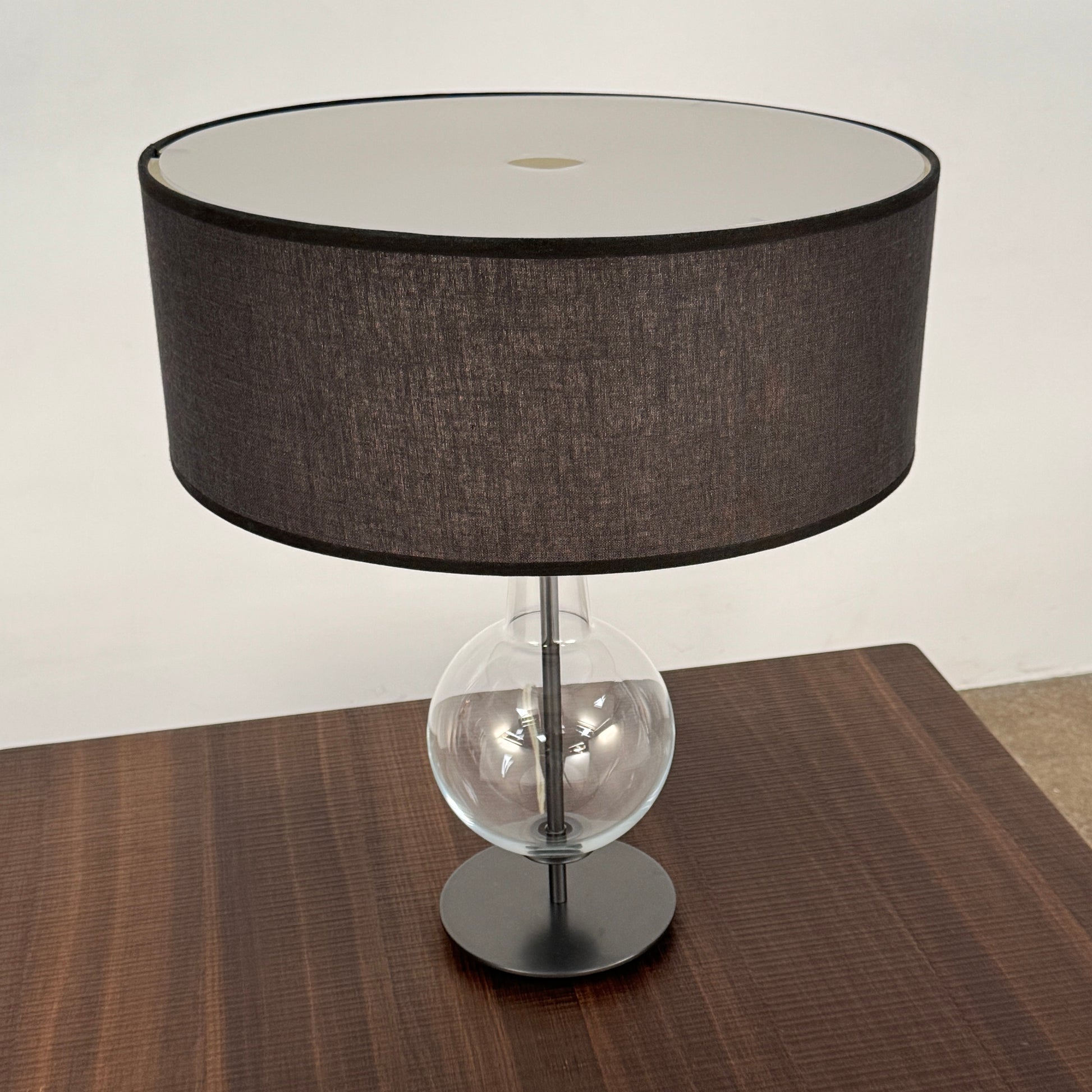The Penta Classic Desir Table Lamp by Penta features a hand-blown glass base and a large cylindrical black lampshade, showcased against a plain white background.