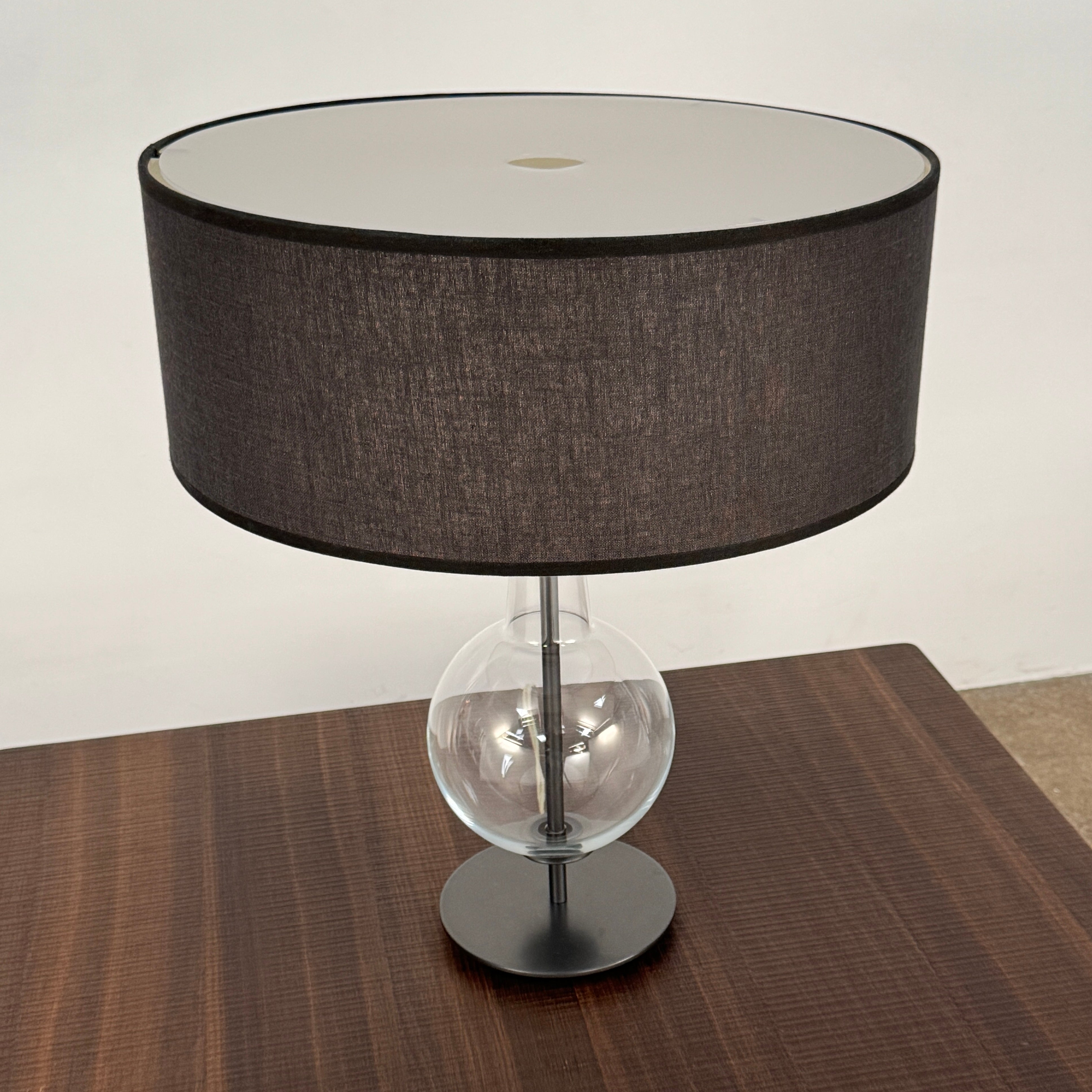 The Penta Classic Desir Table Lamp by Penta features a hand-blown glass base and a large cylindrical black lampshade, showcased against a plain white background.