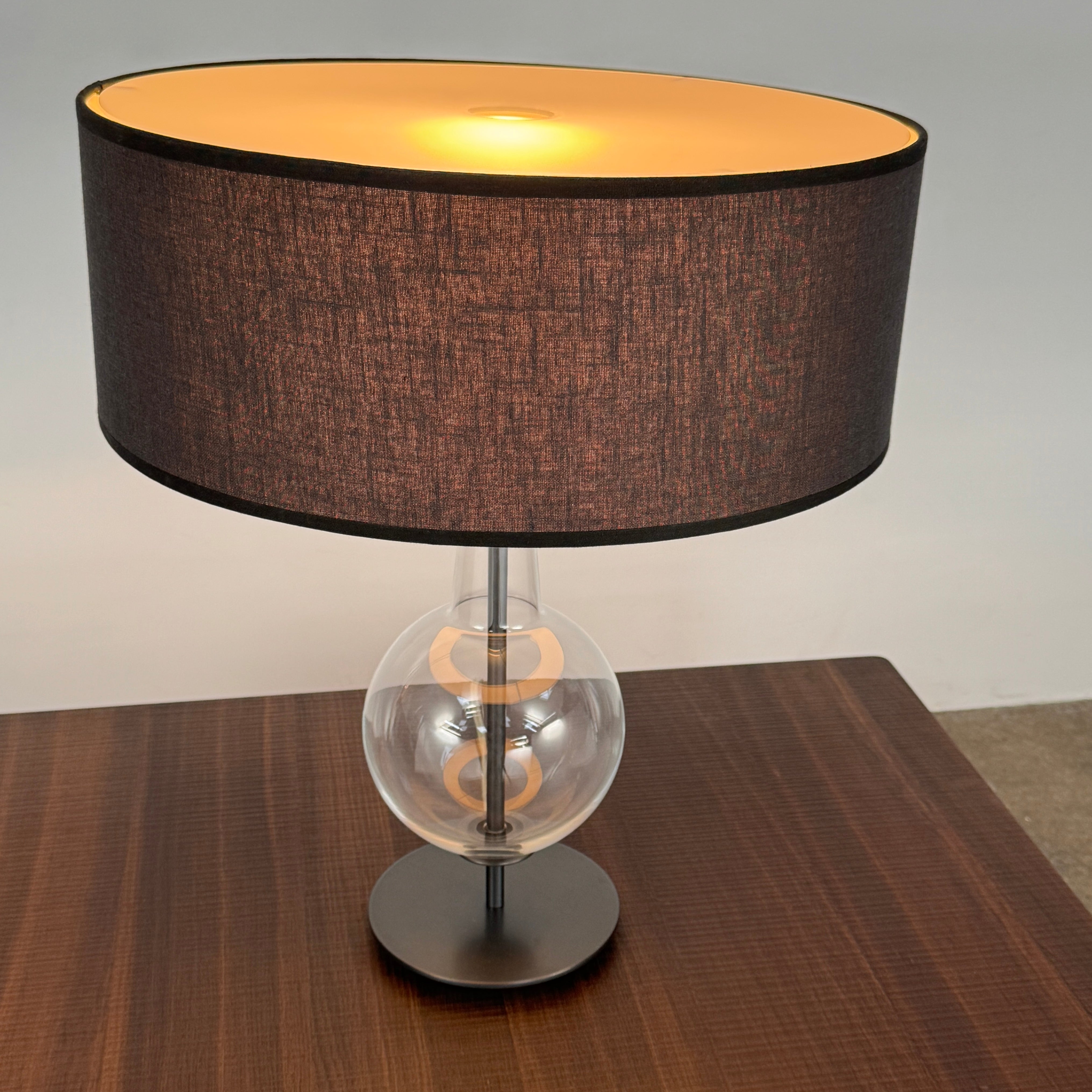 The Penta Classic Desir Table Lamp by Penta features a hand-blown glass base and a large cylindrical black lampshade, showcased against a plain white background.