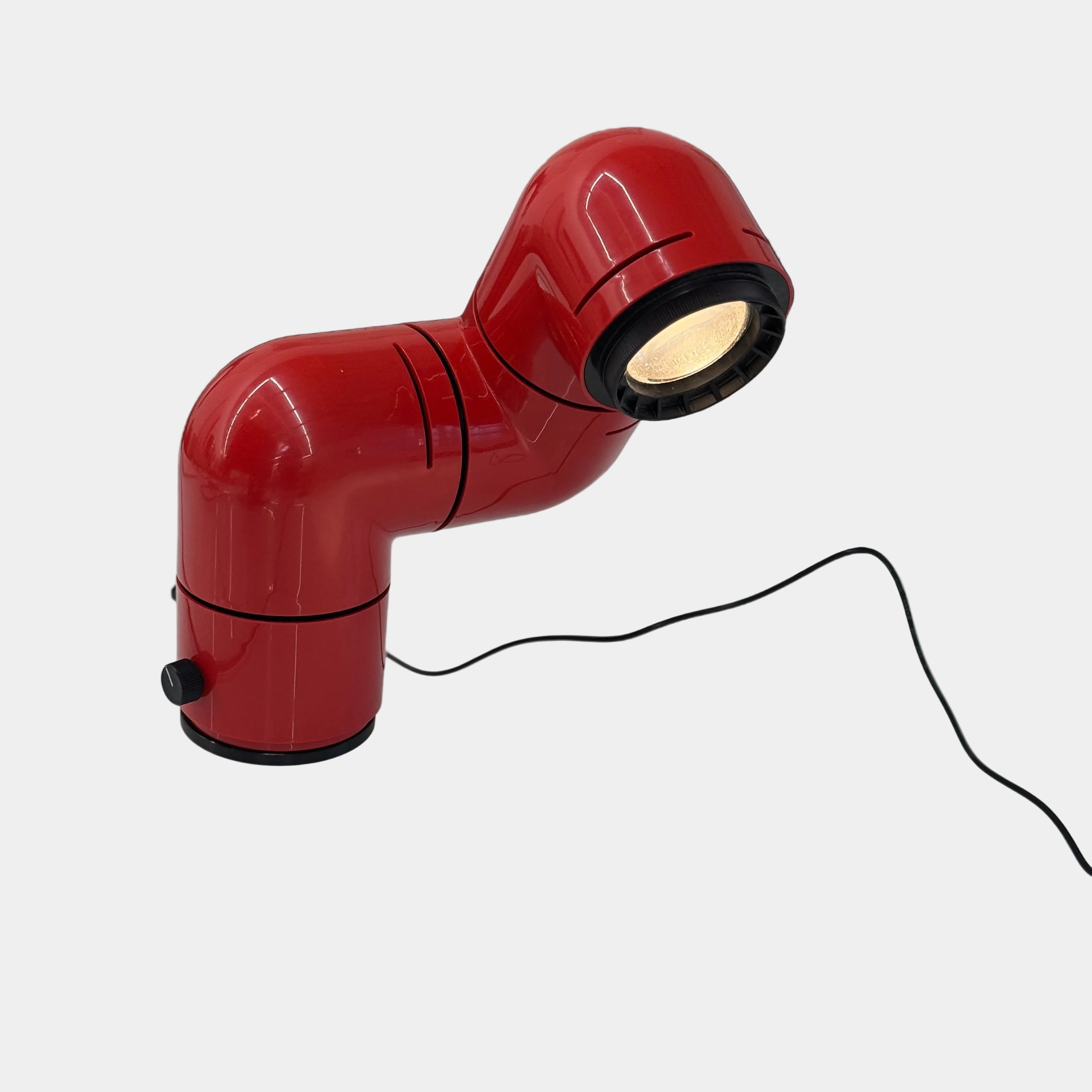 A Santa & Cole Tatu Red Table Lamp with a pipe-shaped design and spotlight head rests on a white surface, plugged in with its black cord.