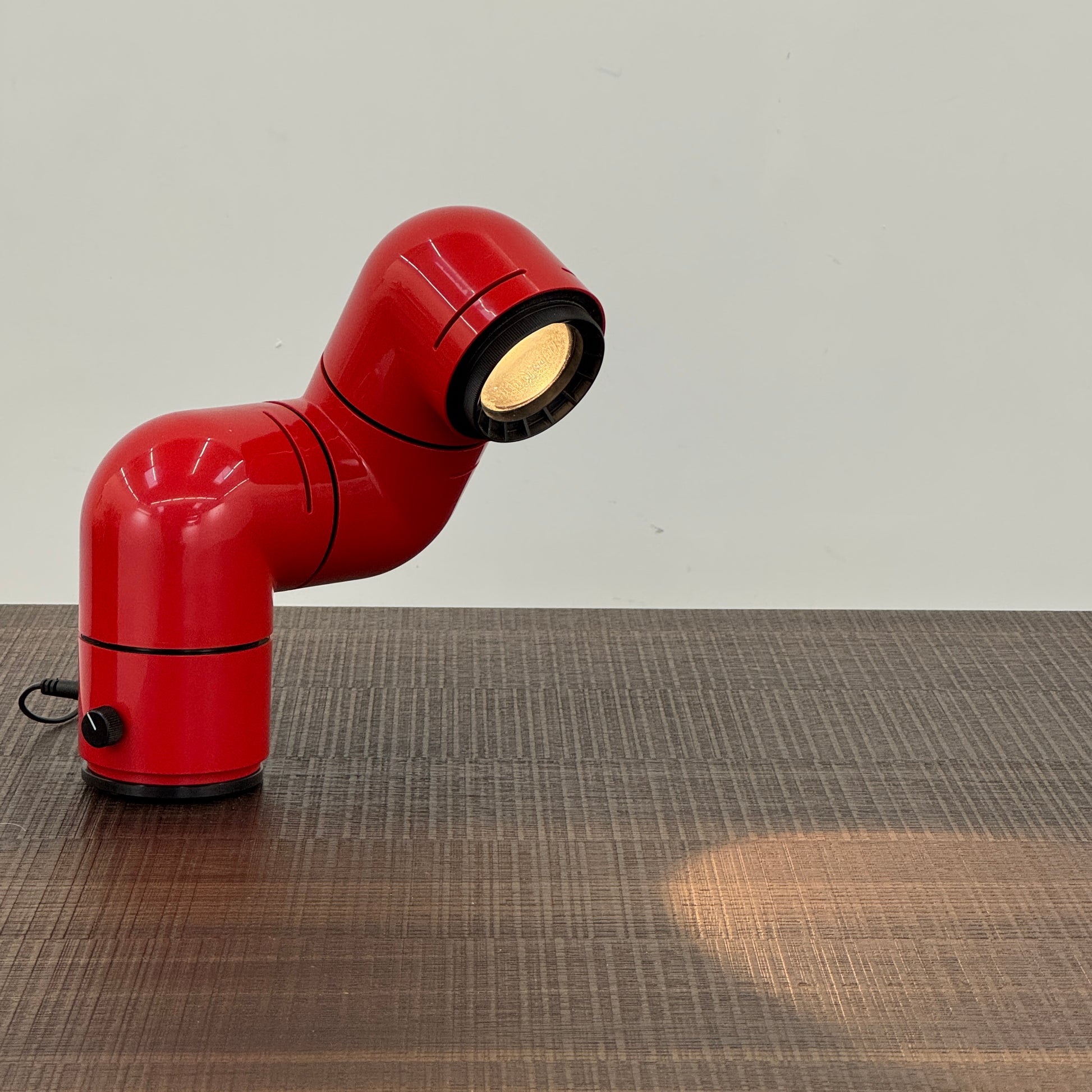 A Santa & Cole Tatu Red Table Lamp with a pipe-shaped design and spotlight head rests on a white surface, plugged in with its black cord.