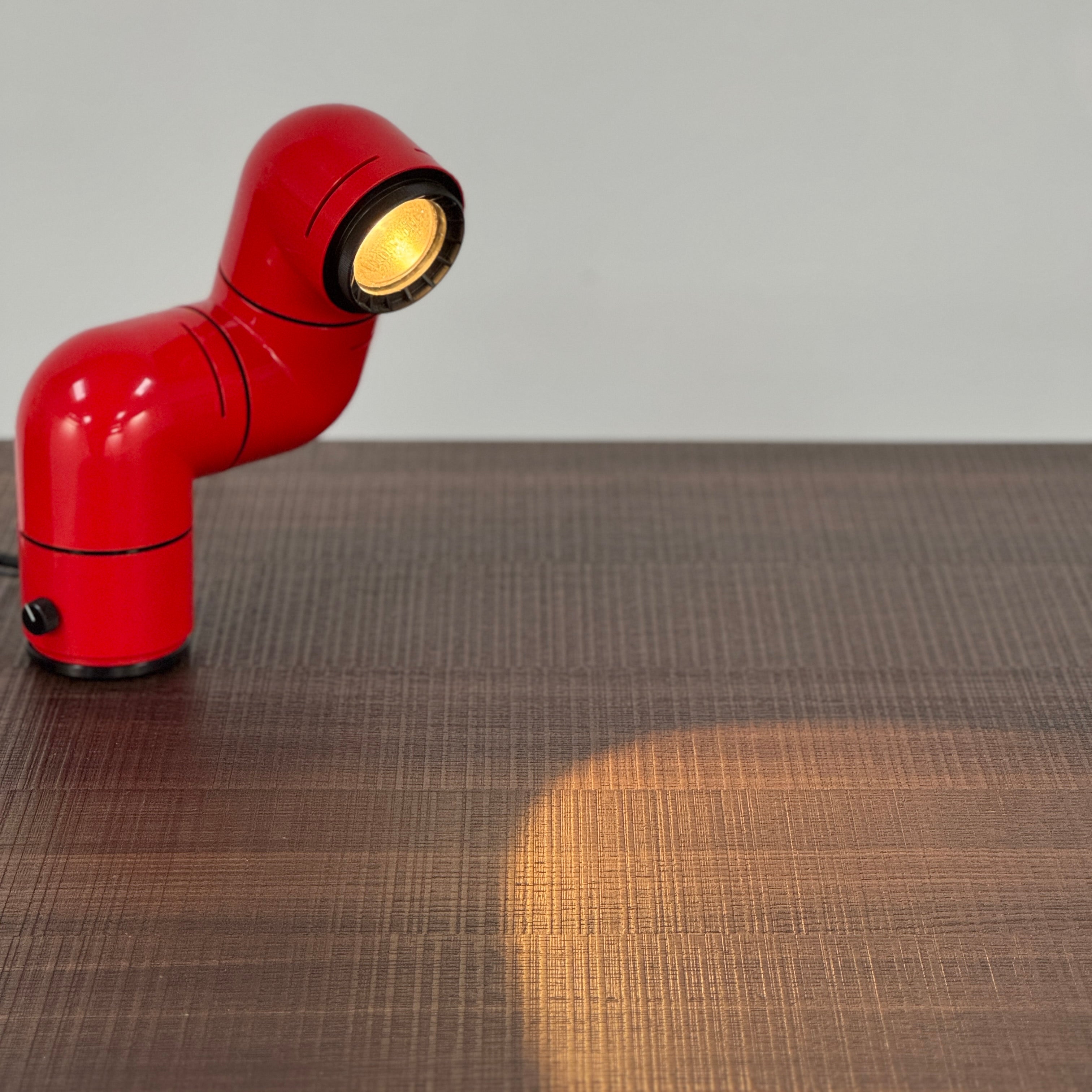 A Santa & Cole Tatu Red Table Lamp with a pipe-shaped design and spotlight head rests on a white surface, plugged in with its black cord.
