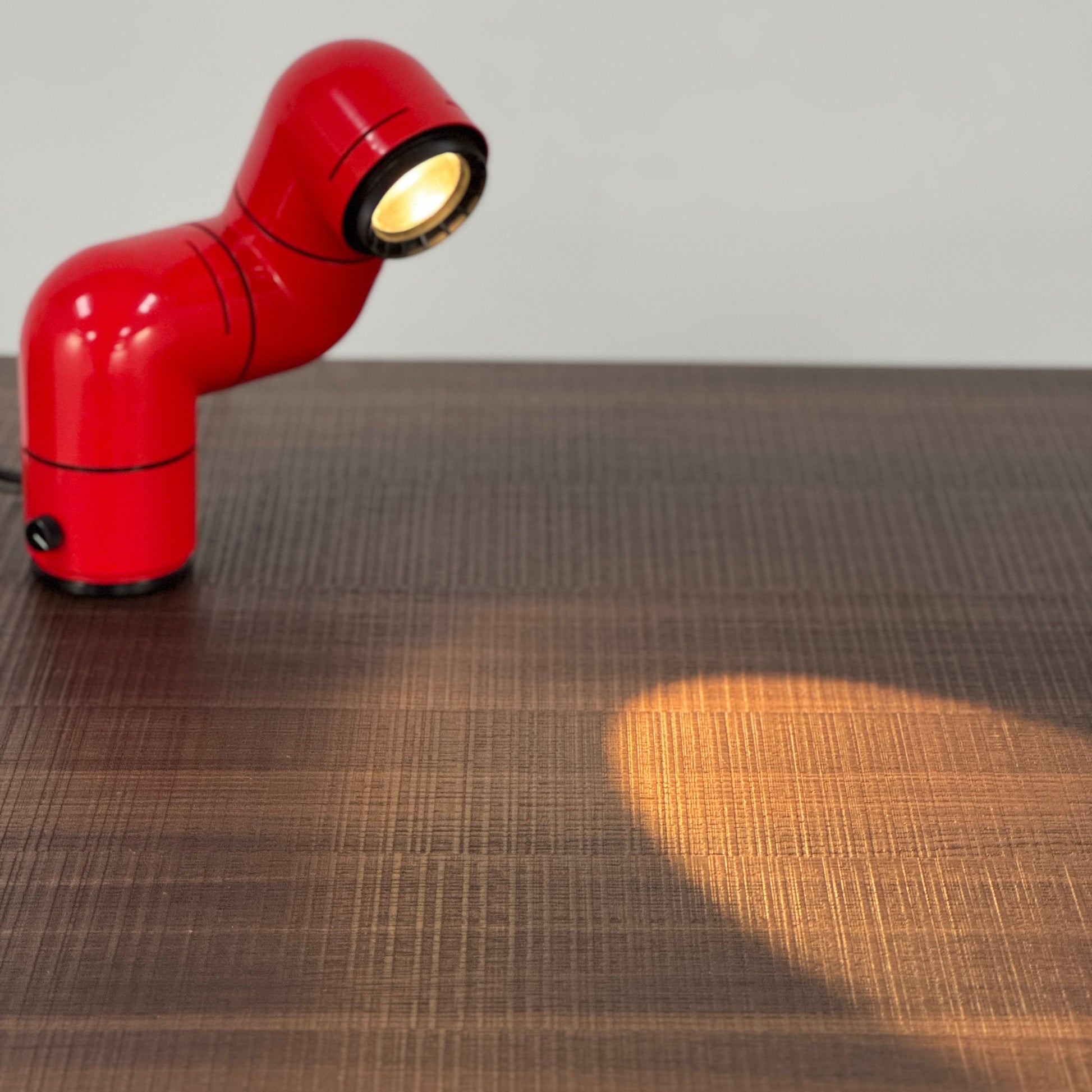 A Santa & Cole Tatu Red Table Lamp with a pipe-shaped design and spotlight head rests on a white surface, plugged in with its black cord.