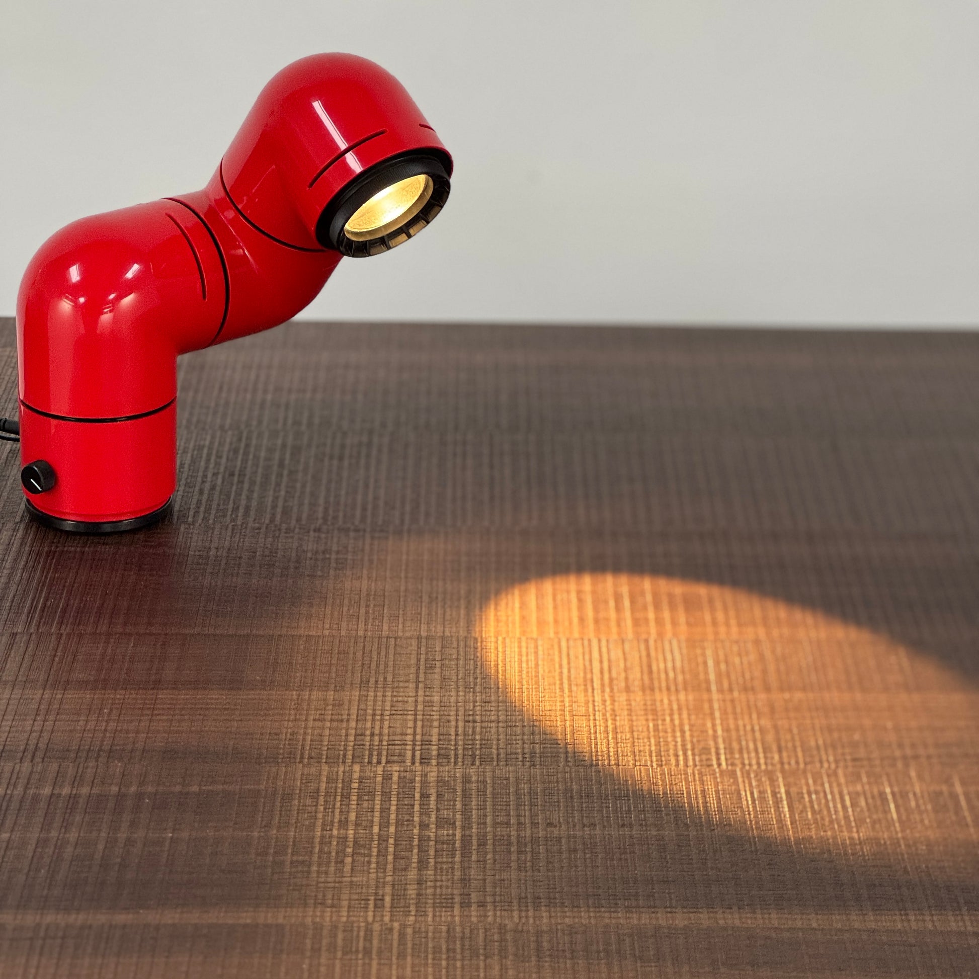A Santa & Cole Tatu Red Table Lamp with a pipe-shaped design and spotlight head rests on a white surface, plugged in with its black cord.