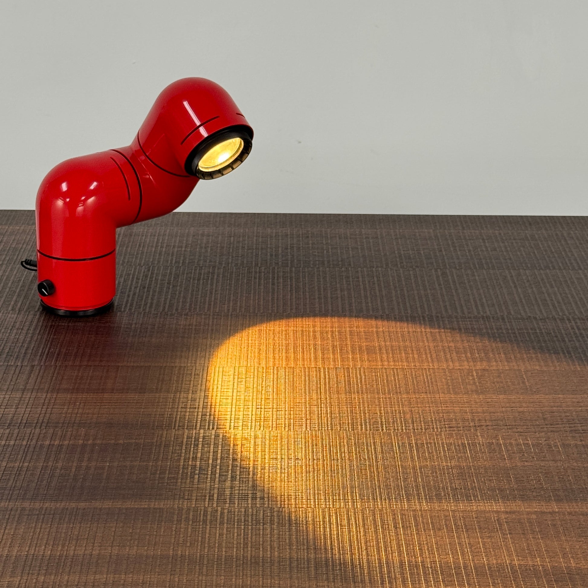 A Santa & Cole Tatu Red Table Lamp with a pipe-shaped design and spotlight head rests on a white surface, plugged in with its black cord.