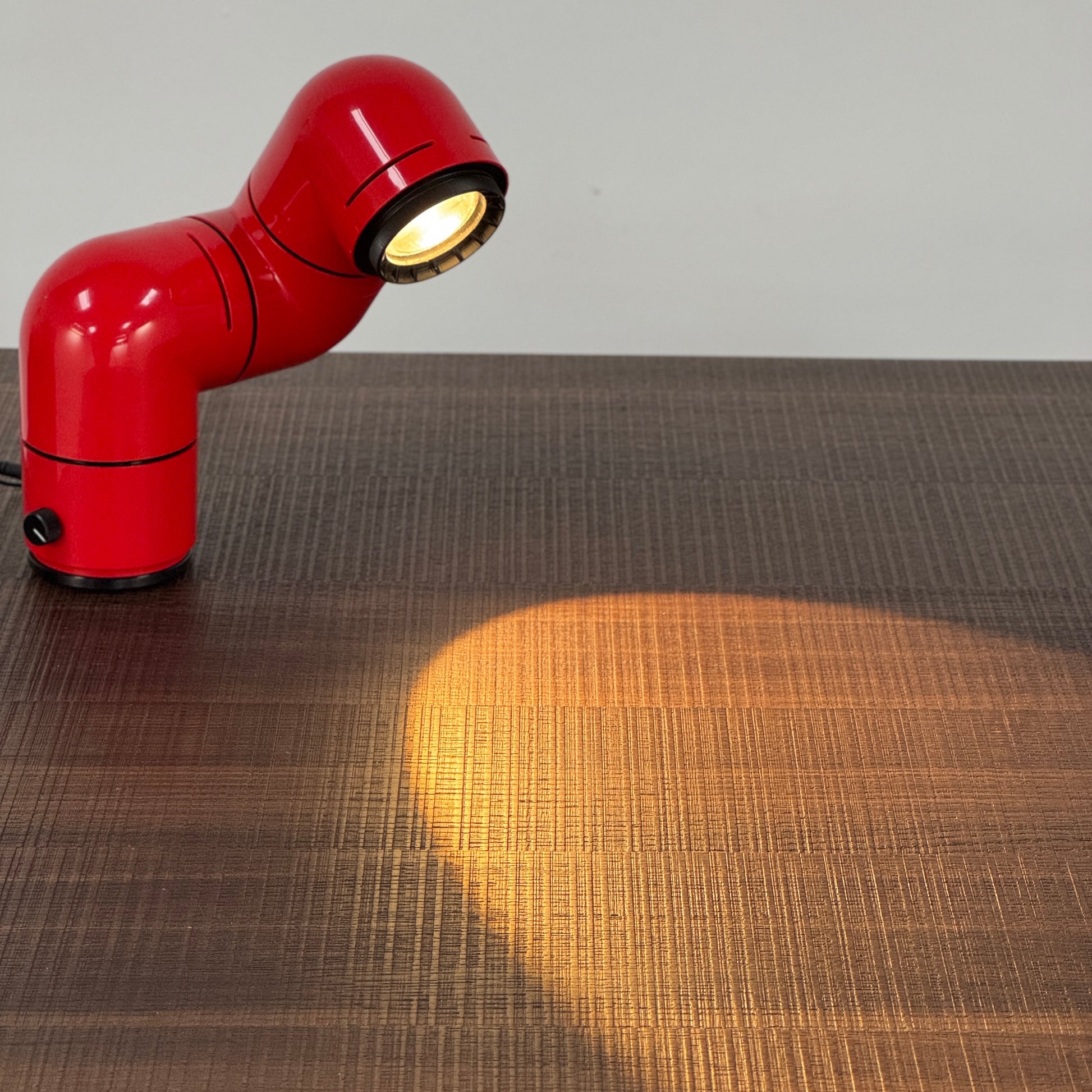 A Santa & Cole Tatu Red Table Lamp with a pipe-shaped design and spotlight head rests on a white surface, plugged in with its black cord.