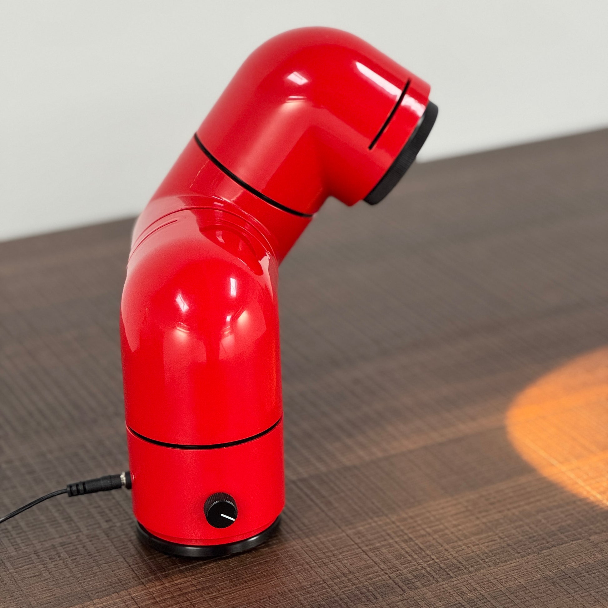 A Santa & Cole Tatu Red Table Lamp with a pipe-shaped design and spotlight head rests on a white surface, plugged in with its black cord.