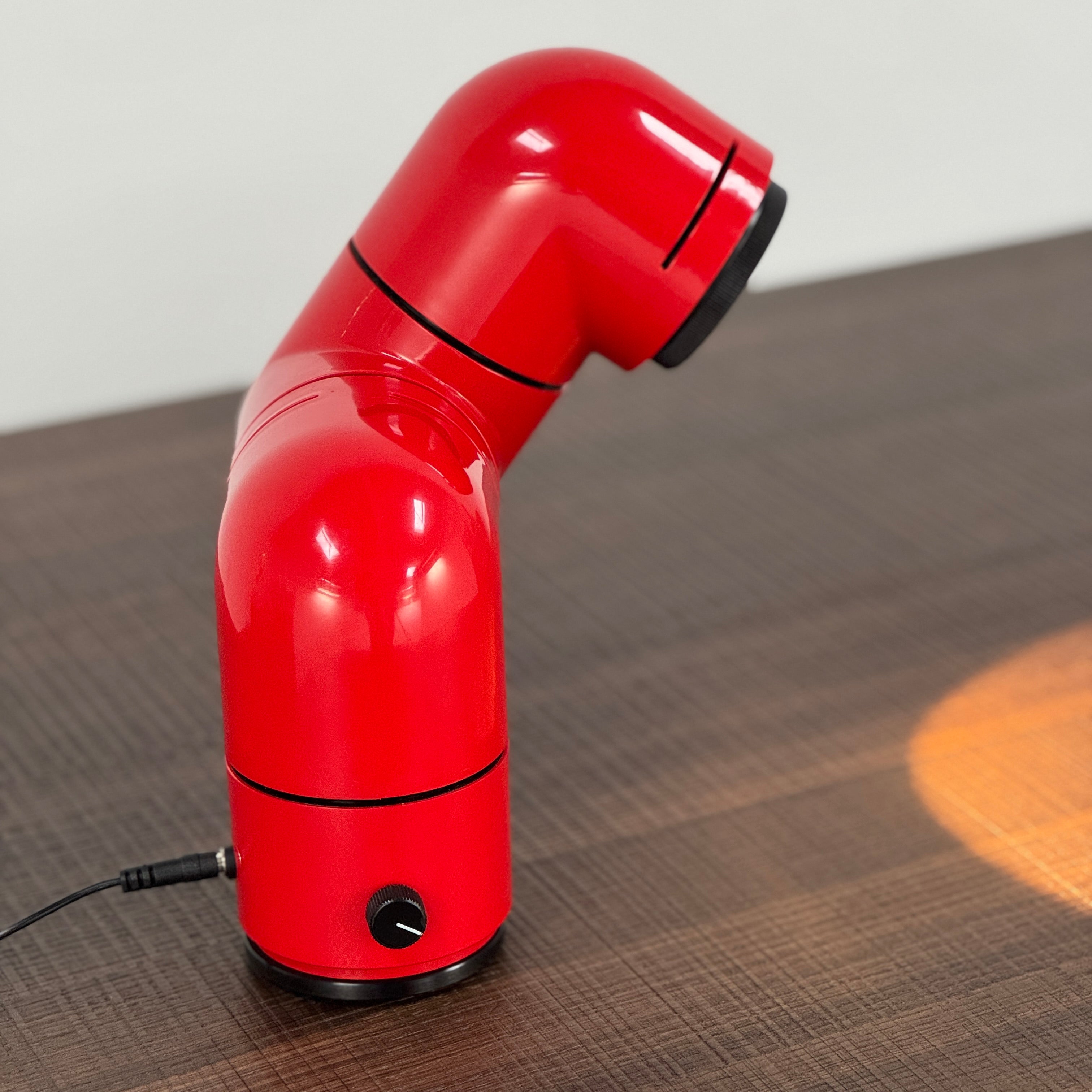 A Santa & Cole Tatu Red Table Lamp with a pipe-shaped design and spotlight head rests on a white surface, plugged in with its black cord.