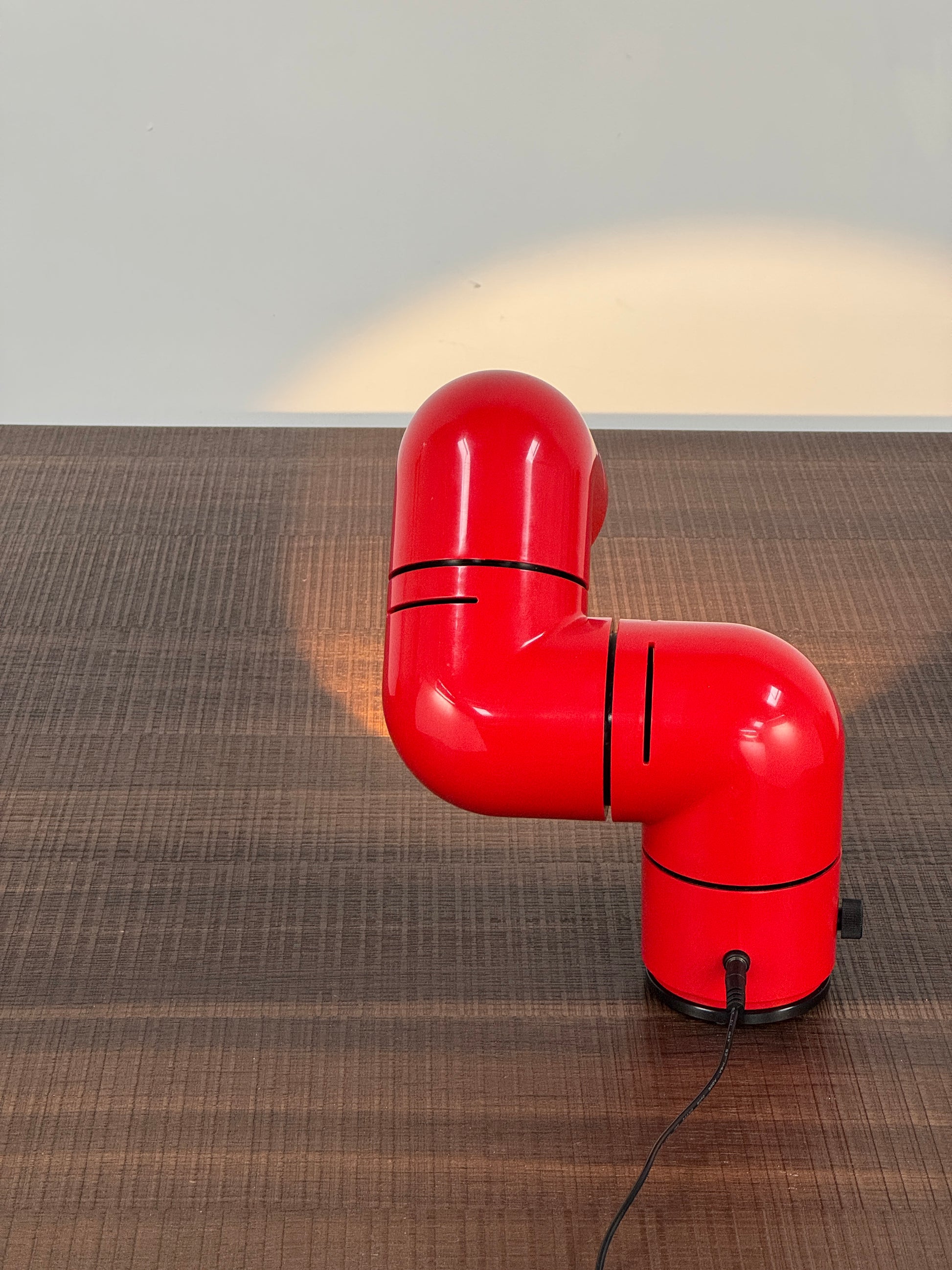 A Santa & Cole Tatu Red Table Lamp with a pipe-shaped design and spotlight head rests on a white surface, plugged in with its black cord.