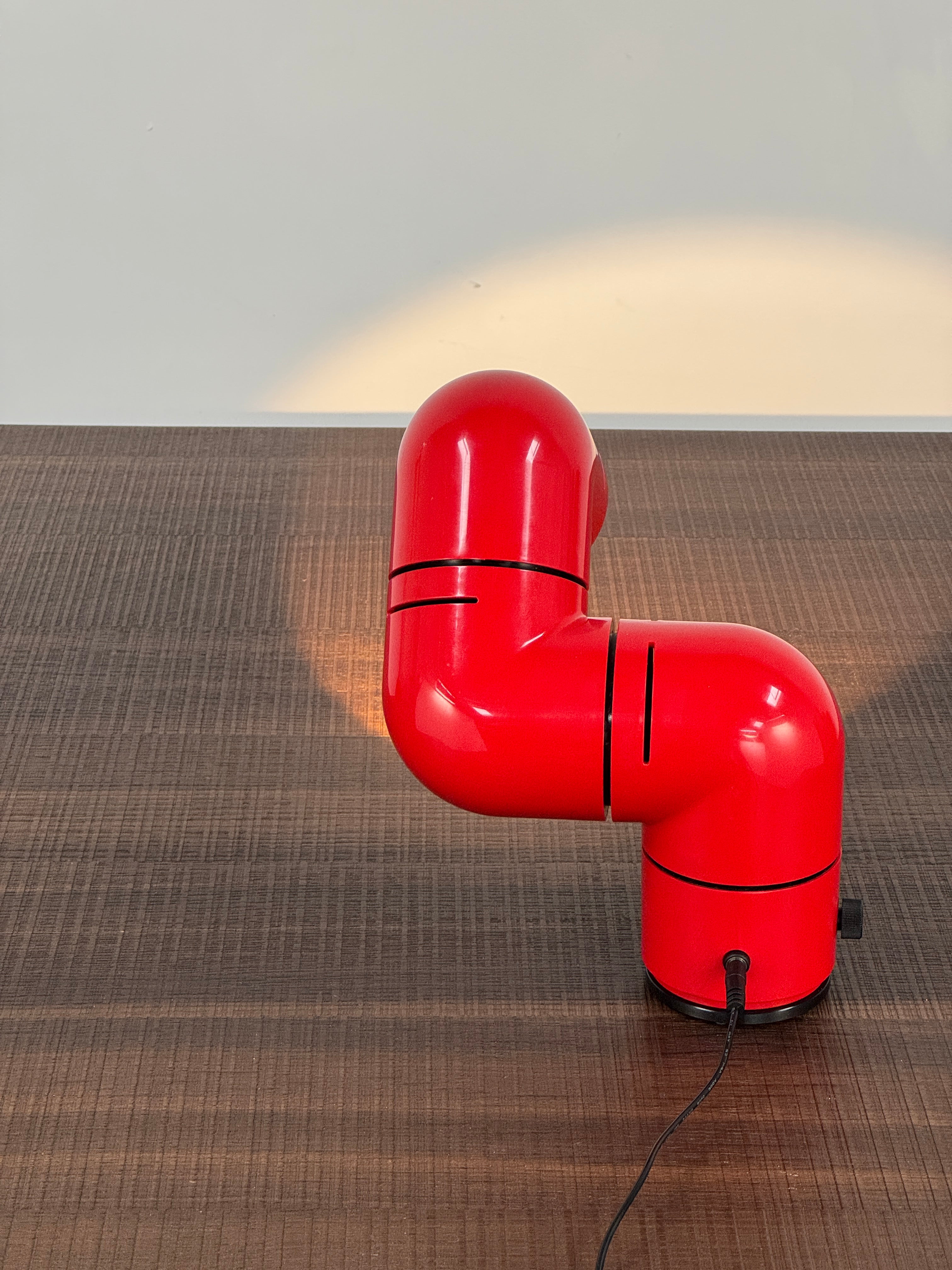 A Santa & Cole Tatu Red Table Lamp with a pipe-shaped design and spotlight head rests on a white surface, plugged in with its black cord.