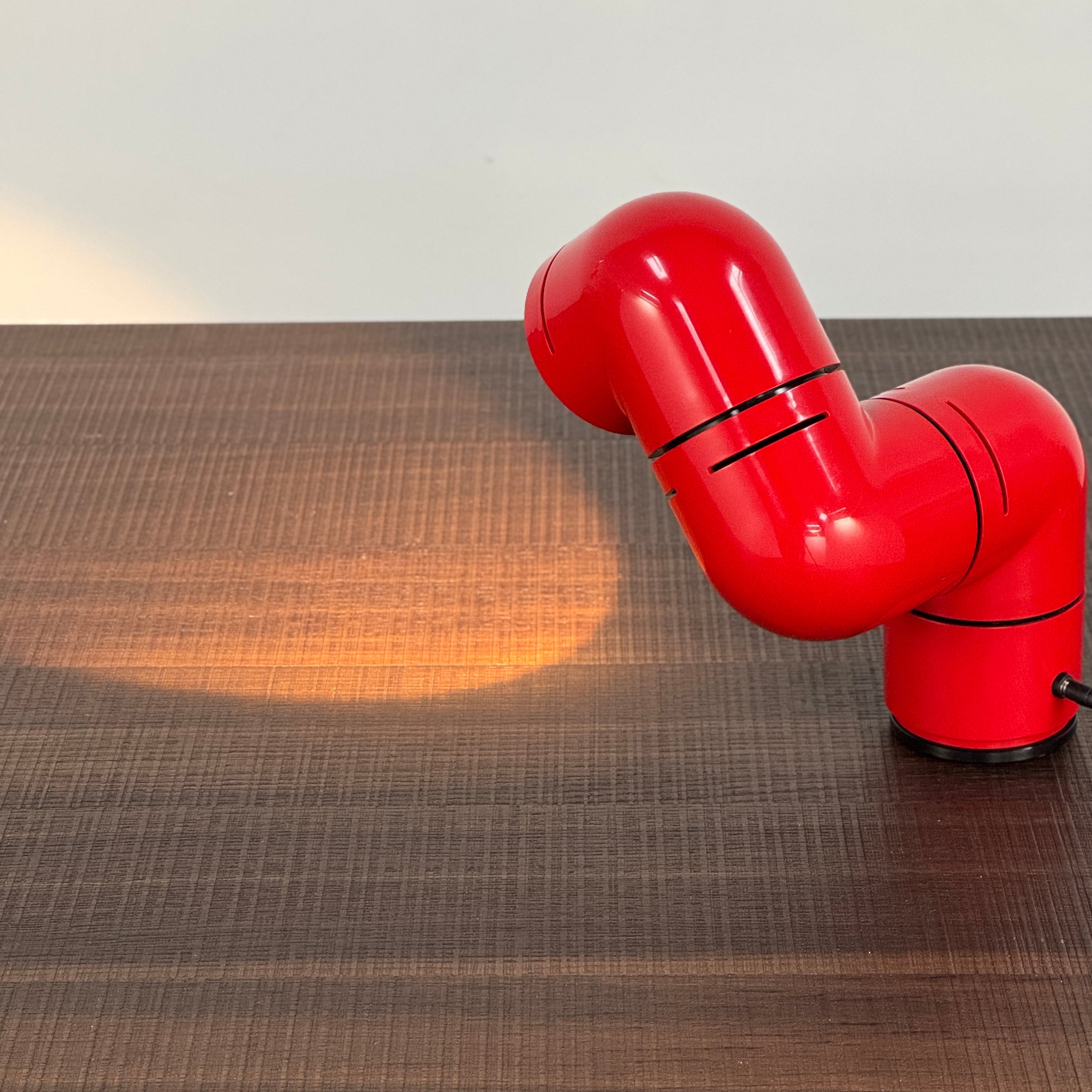 A Santa & Cole Tatu Red Table Lamp with a pipe-shaped design and spotlight head rests on a white surface, plugged in with its black cord.