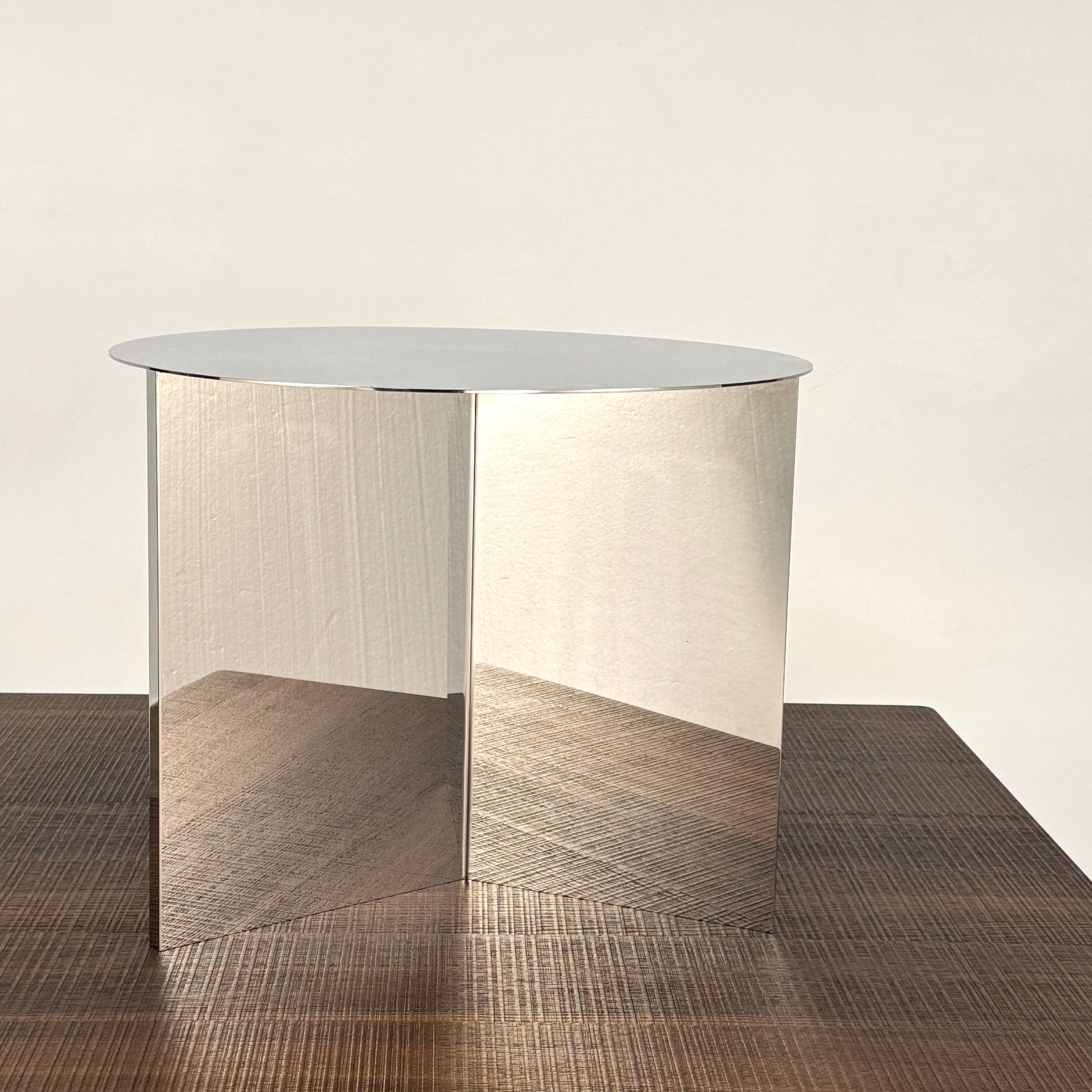 The Hay Slit Side Table by Hay features a round, reflective design with mirrored sides and is shown atop a dark wooden surface against a plain white wall.