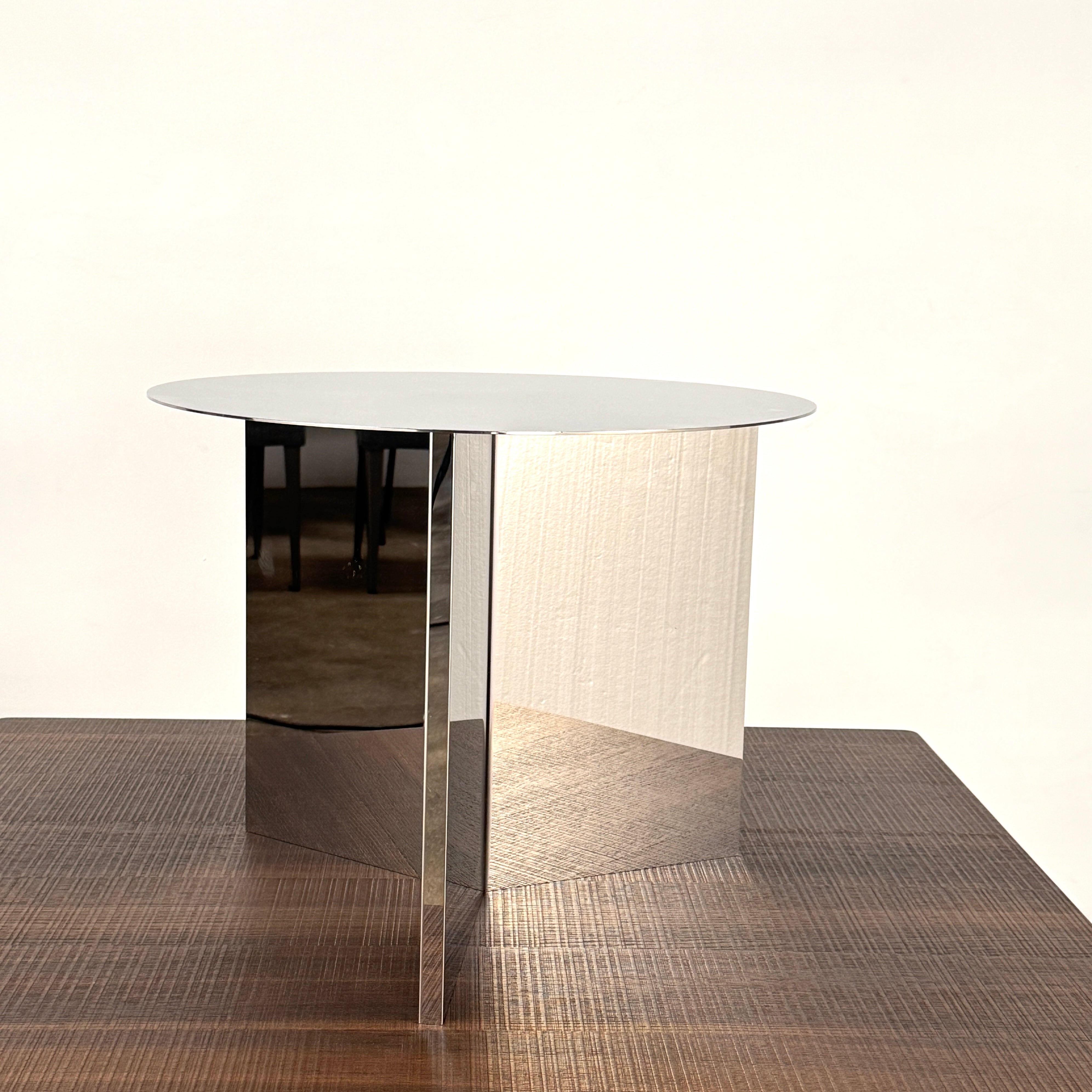 The Hay Slit Side Table by Hay features a round, reflective design with mirrored sides and is shown atop a dark wooden surface against a plain white wall.