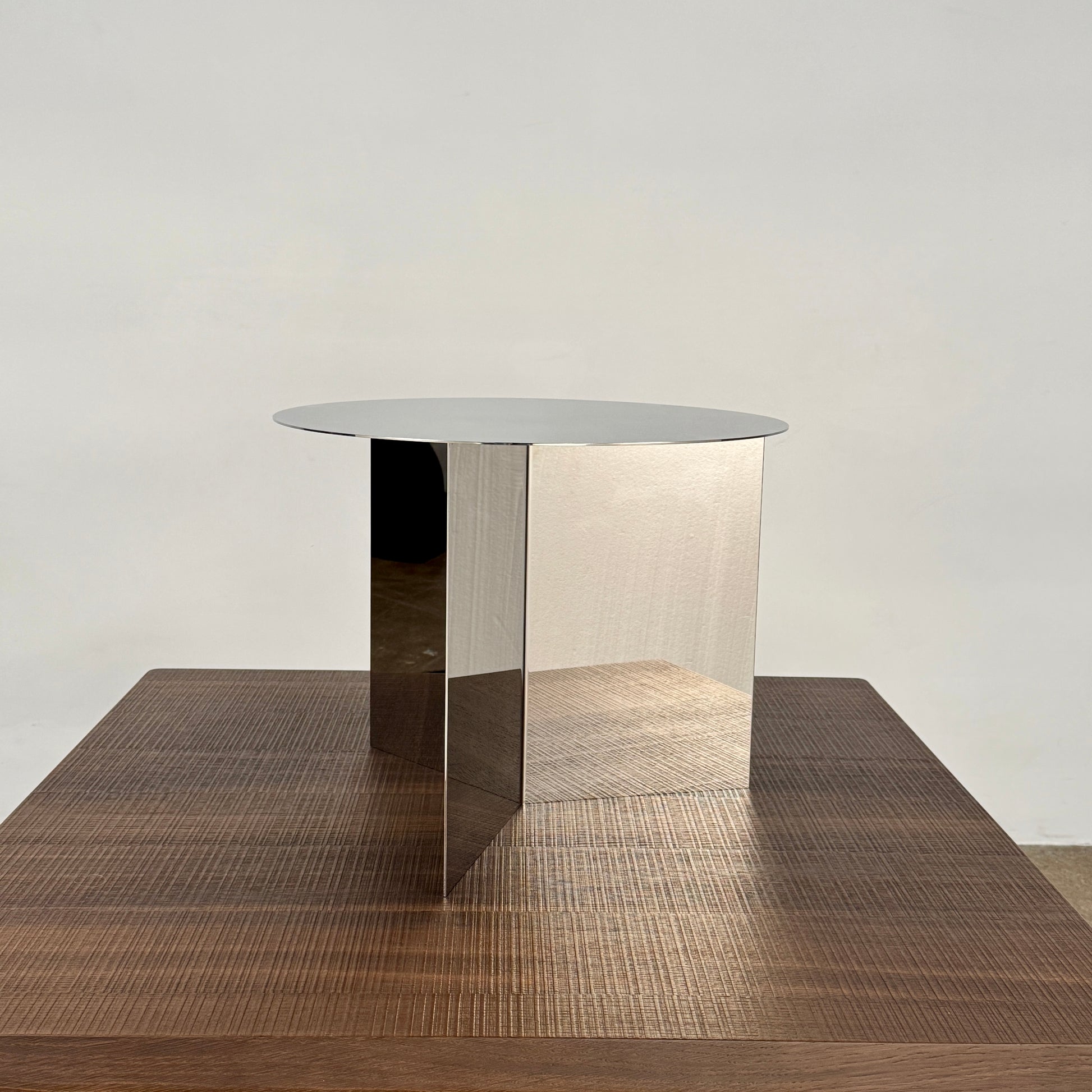 The Hay Slit Side Table by Hay features a round, reflective design with mirrored sides and is shown atop a dark wooden surface against a plain white wall.