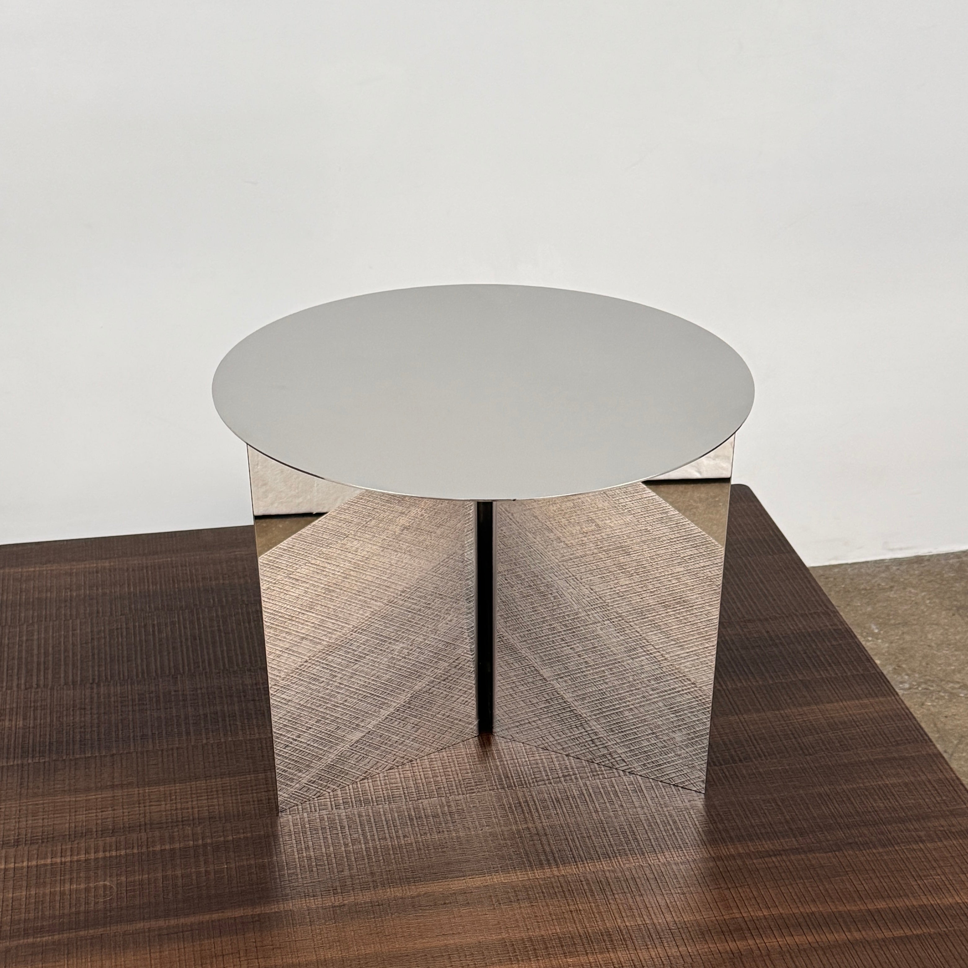 The Hay Slit Side Table by Hay features a round, reflective design with mirrored sides and is shown atop a dark wooden surface against a plain white wall.