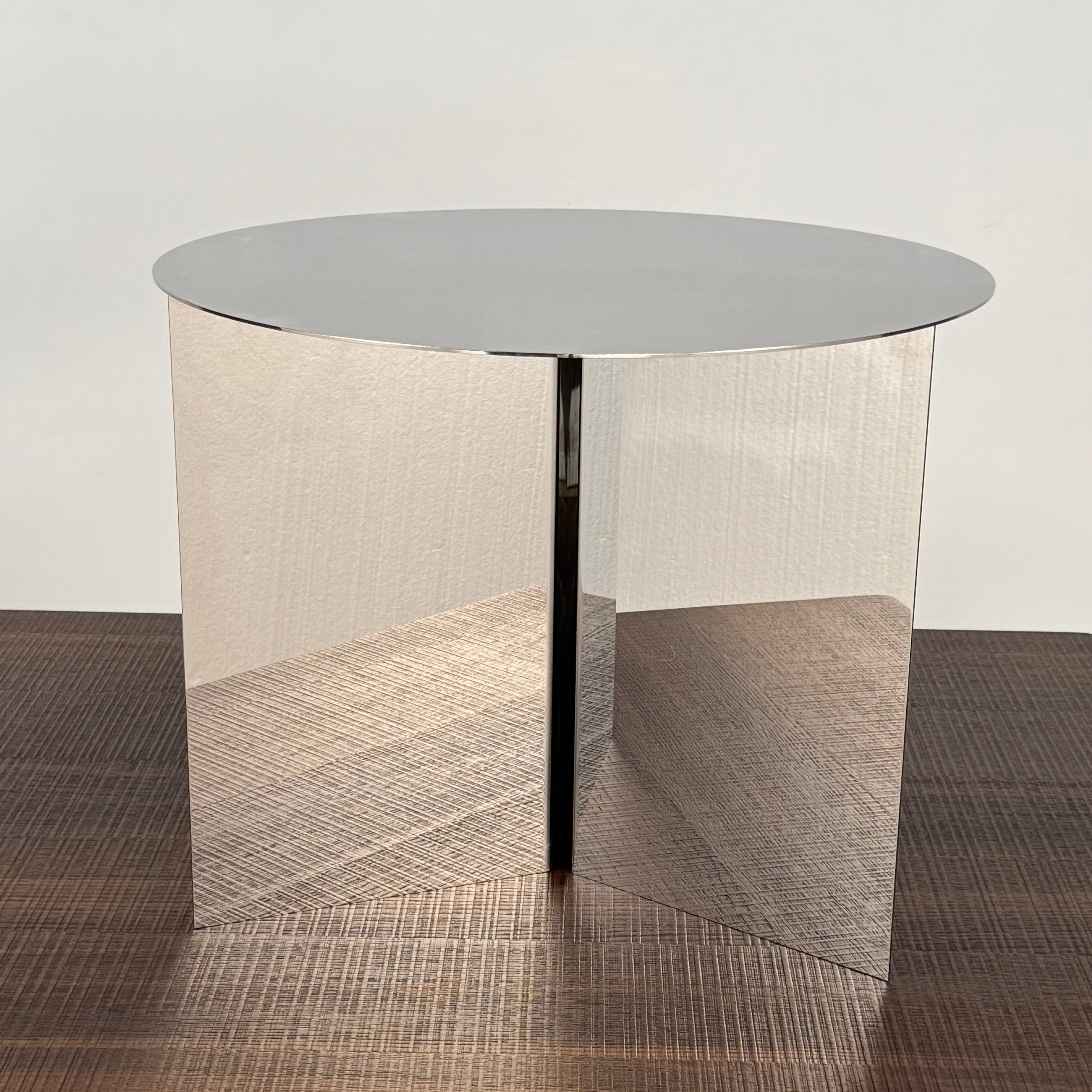 The Hay Slit Side Table by Hay features a round, reflective design with mirrored sides and is shown atop a dark wooden surface against a plain white wall.