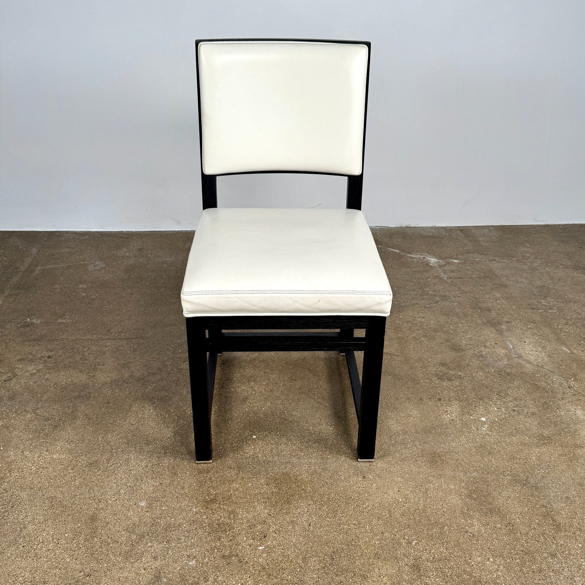 A Maxalto Teti Dining Chair from a set of 6, featuring a black frame with white upholstered seat and backrest, stands on a brown concrete floor against a plain light gray wall.