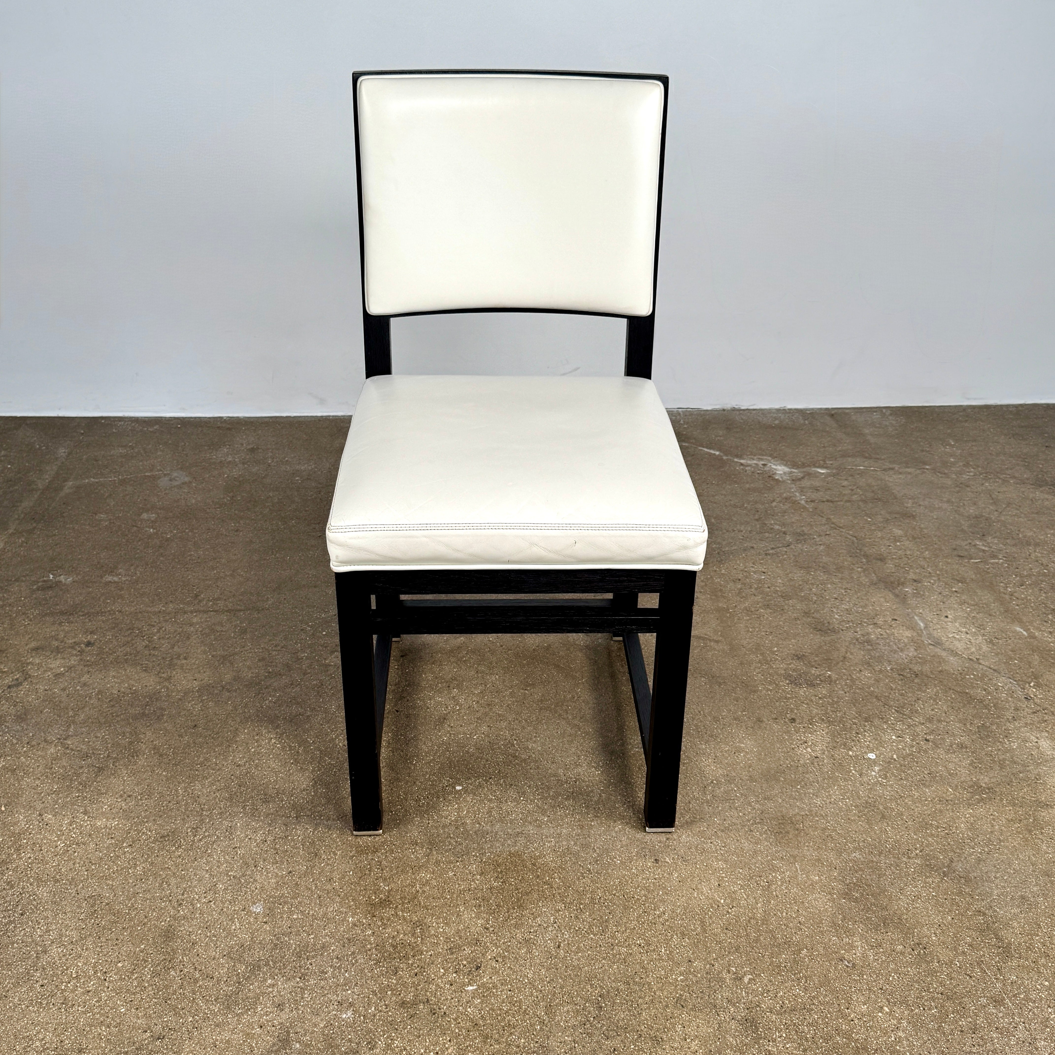A Maxalto Teti Dining Chair from a set of 6, featuring a black frame with white upholstered seat and backrest, stands on a brown concrete floor against a plain light gray wall.