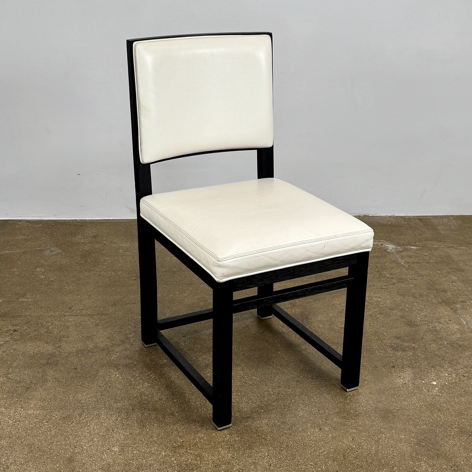 A Maxalto Teti Dining Chair from a set of 6, featuring a black frame with white upholstered seat and backrest, stands on a brown concrete floor against a plain light gray wall.