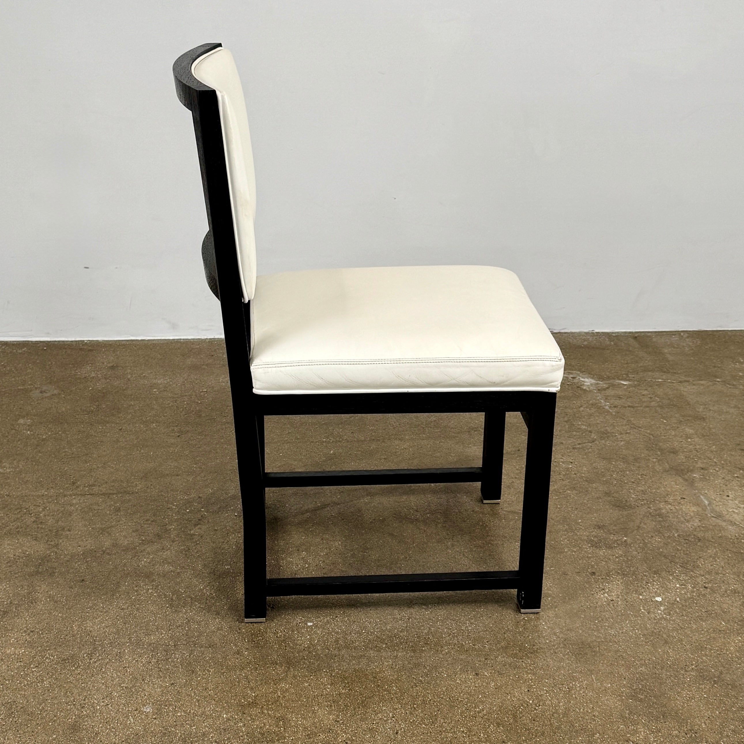 A Maxalto Teti Dining Chair from a set of 6, featuring a black frame with white upholstered seat and backrest, stands on a brown concrete floor against a plain light gray wall.