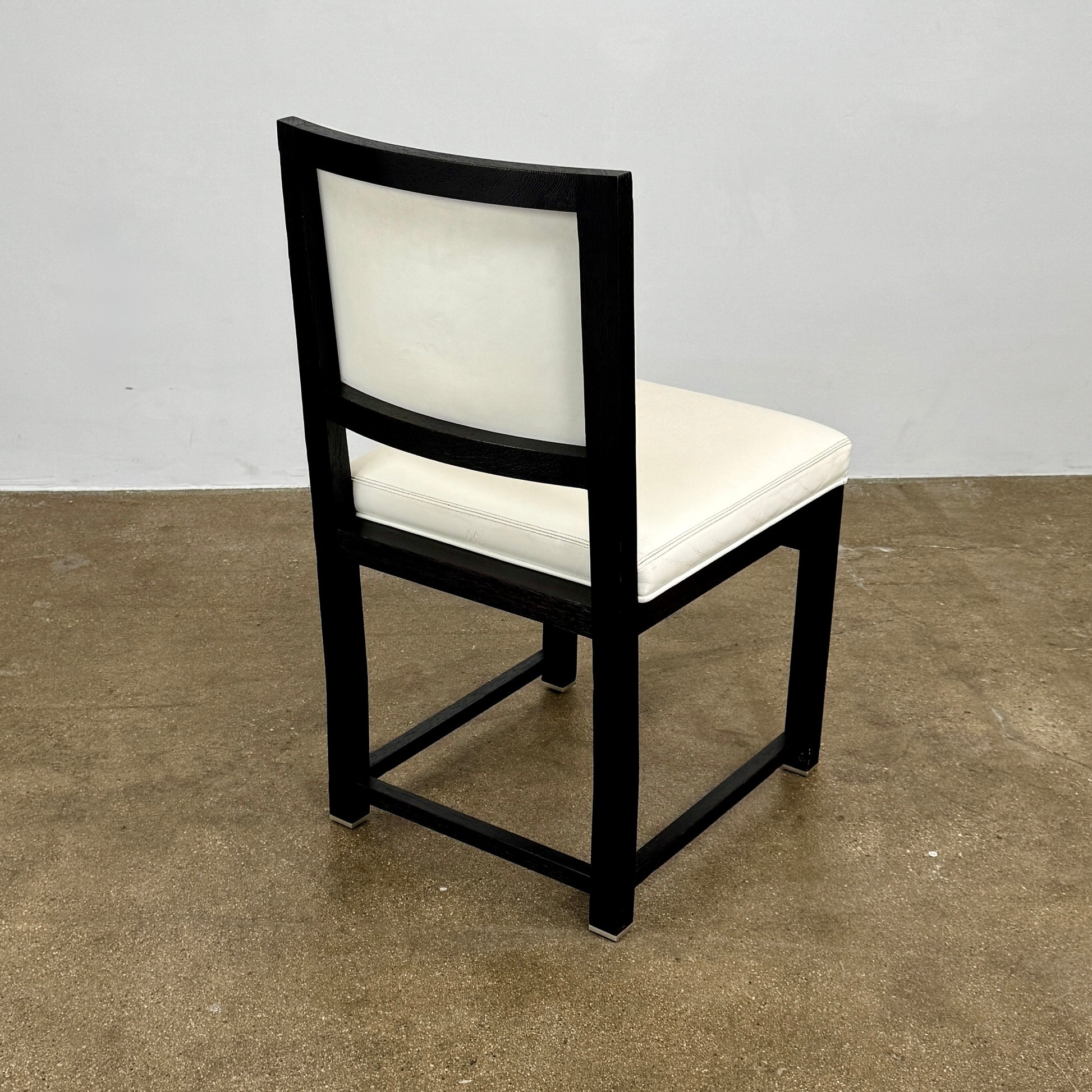 A Maxalto Teti Dining Chair from a set of 6, featuring a black frame with white upholstered seat and backrest, stands on a brown concrete floor against a plain light gray wall.