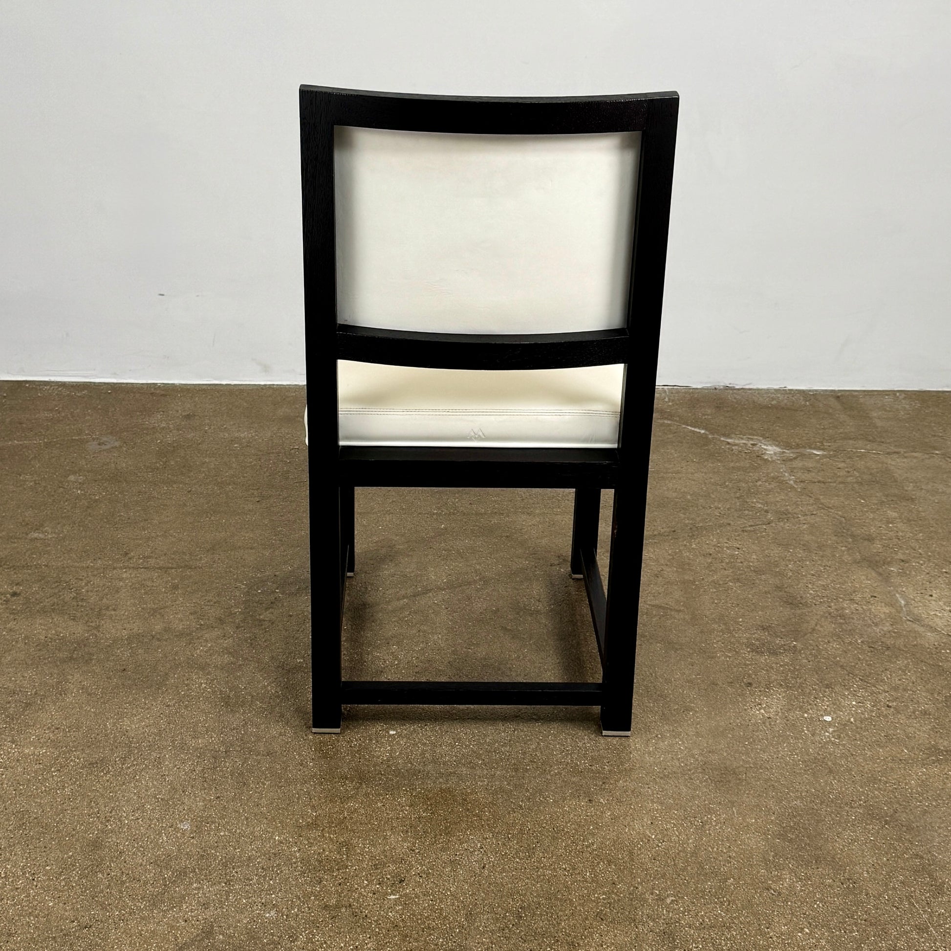 A Maxalto Teti Dining Chair from a set of 6, featuring a black frame with white upholstered seat and backrest, stands on a brown concrete floor against a plain light gray wall.