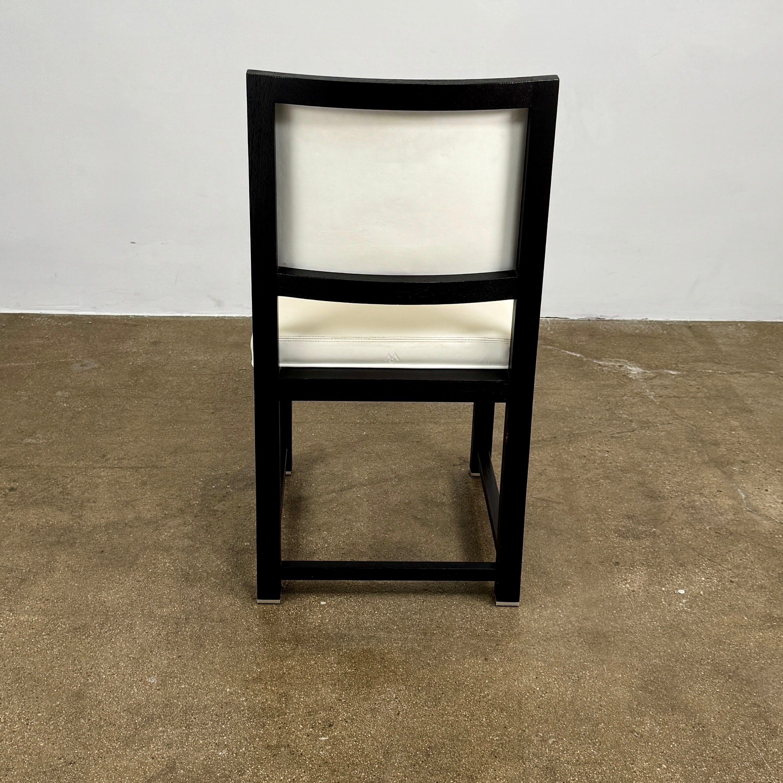 A Maxalto Teti Dining Chair from a set of 6, featuring a black frame with white upholstered seat and backrest, stands on a brown concrete floor against a plain light gray wall.