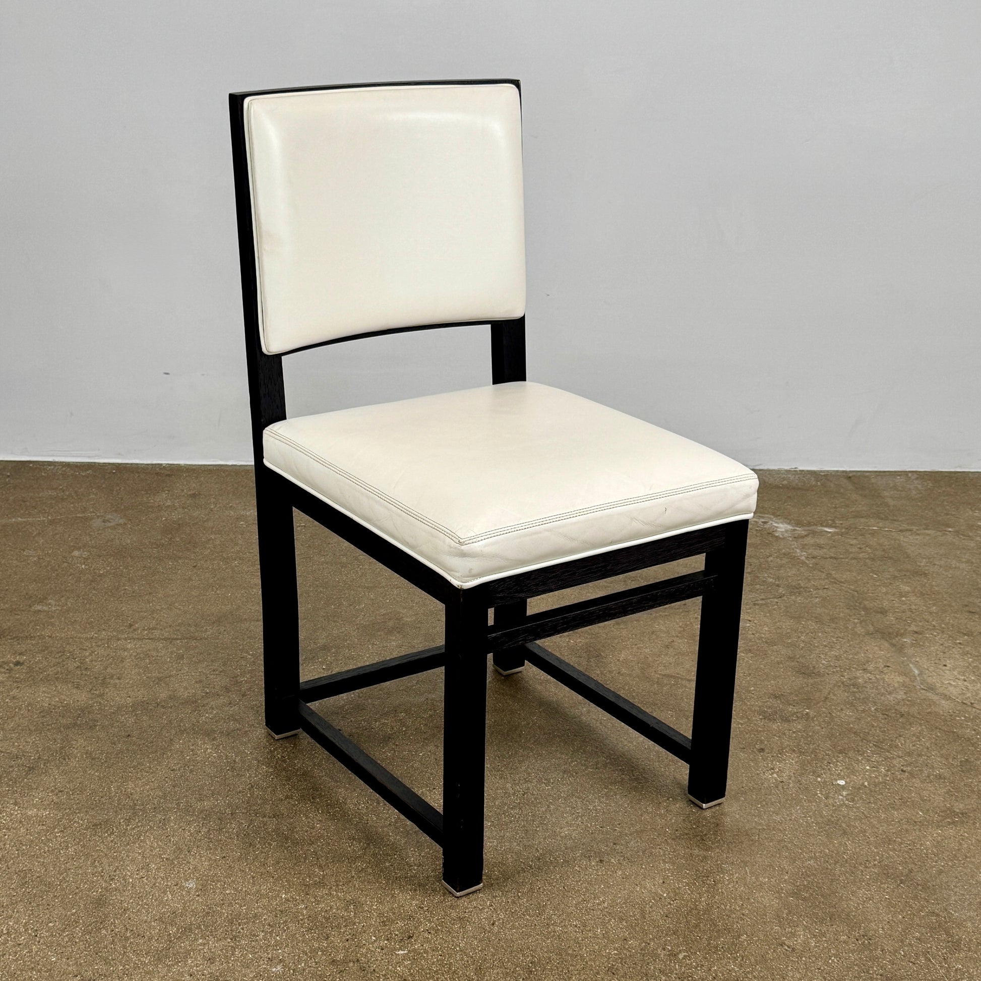 A Maxalto Teti Dining Chair from a set of 6, featuring a black frame with white upholstered seat and backrest, stands on a brown concrete floor against a plain light gray wall.