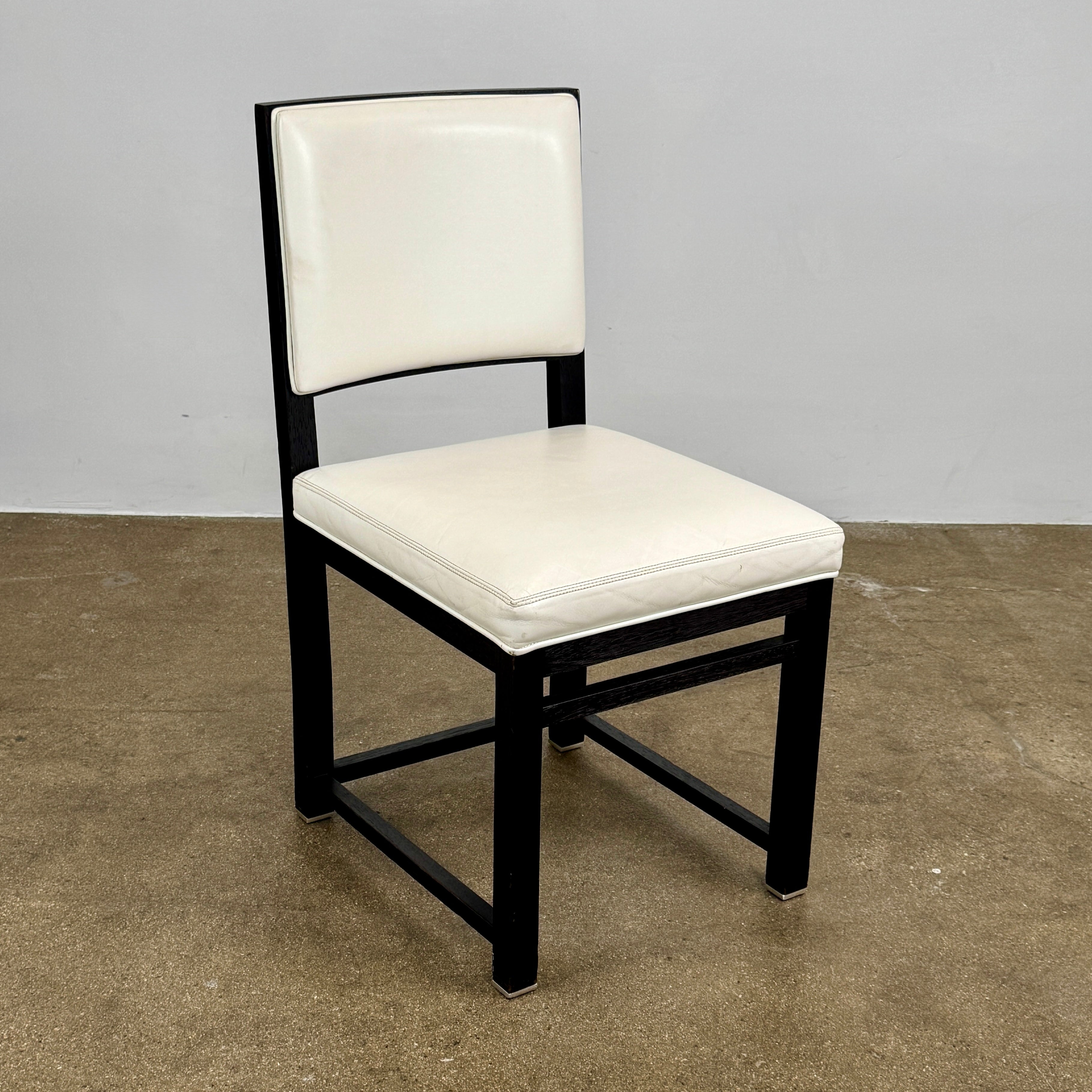 A Maxalto Teti Dining Chair from a set of 6, featuring a black frame with white upholstered seat and backrest, stands on a brown concrete floor against a plain light gray wall.