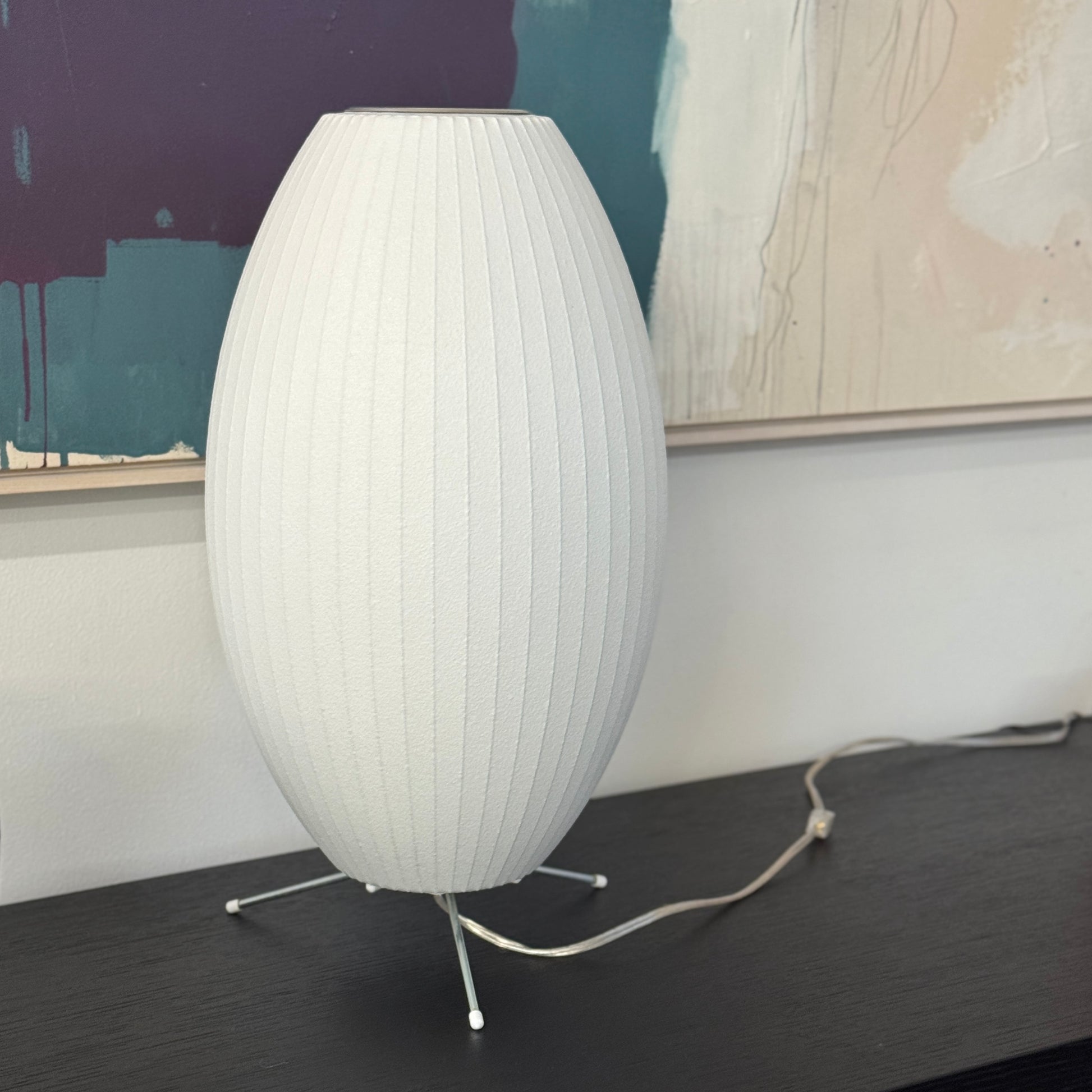 A white, oval-shaped Herman Miller Nelson Cigar Tripod Lamp with vertical ridges rests on a black surface, its cord trailing behind, while an abstract painting hangs in the background.