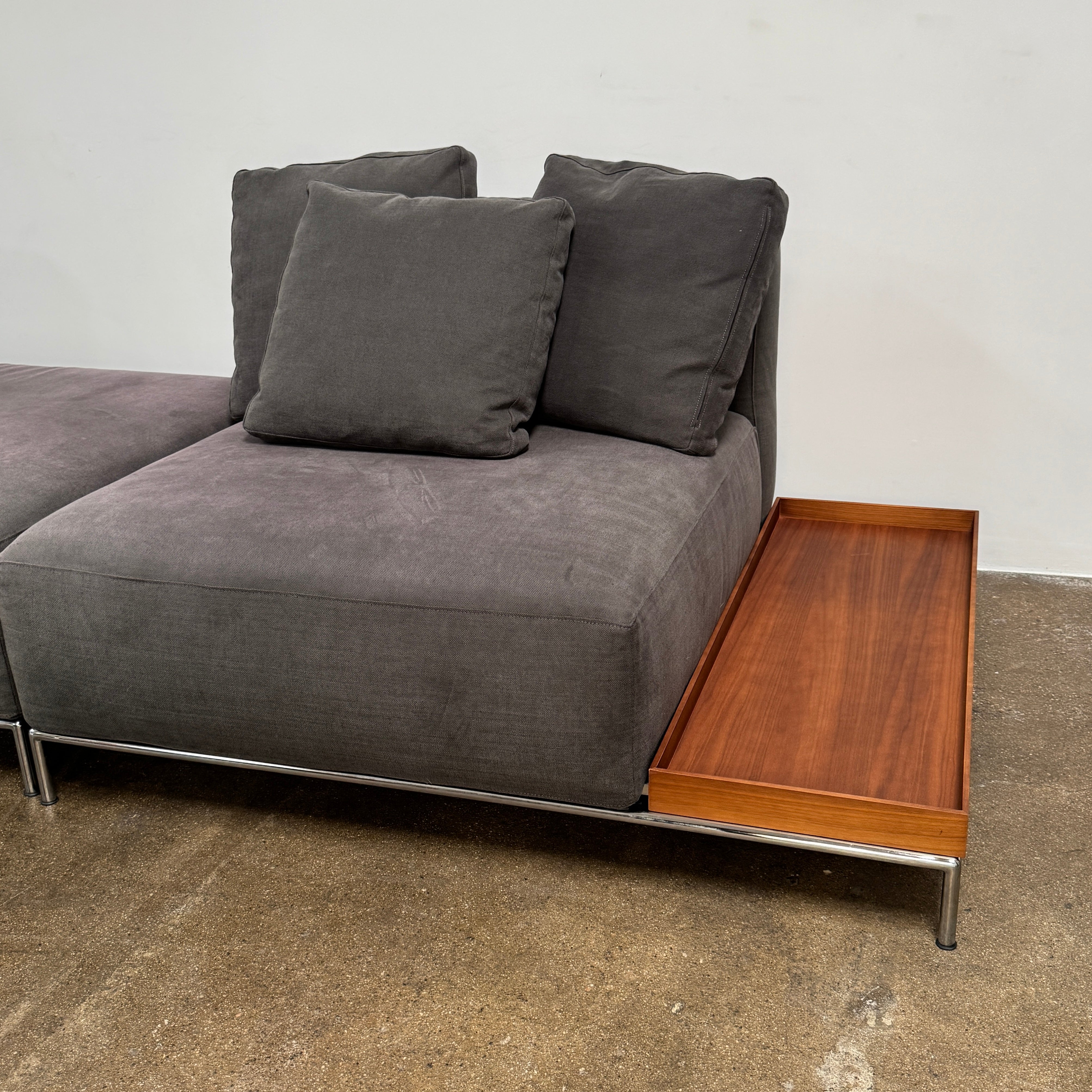 The Cassina Mex Hi Modular Sofa by Cassina features dark gray recycled fiber upholstery, metal legs, multiple cushions, and an integrated wooden side table, shown against a white background.