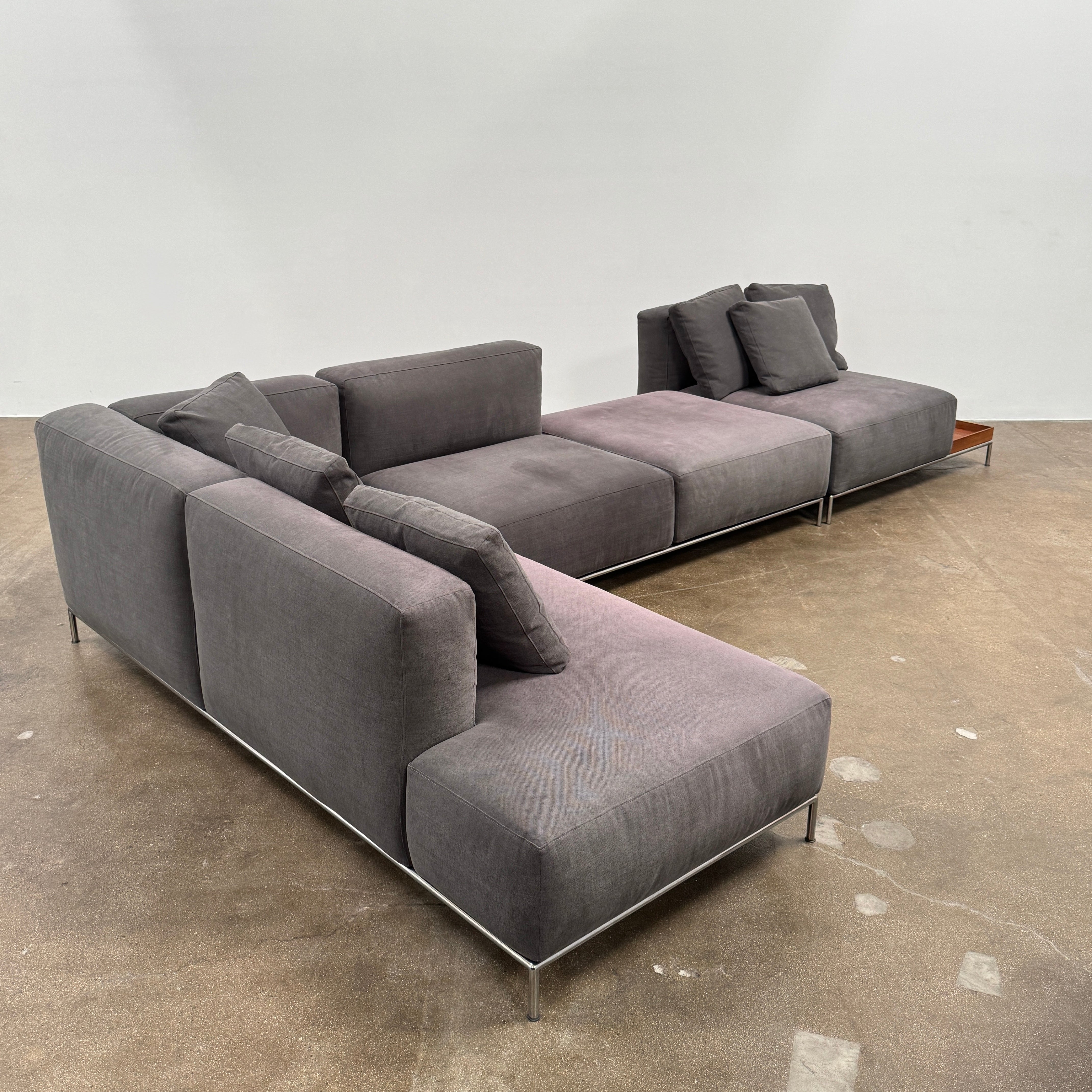 The Cassina Mex Hi Modular Sofa by Cassina features dark gray recycled fiber upholstery, metal legs, multiple cushions, and an integrated wooden side table, shown against a white background.