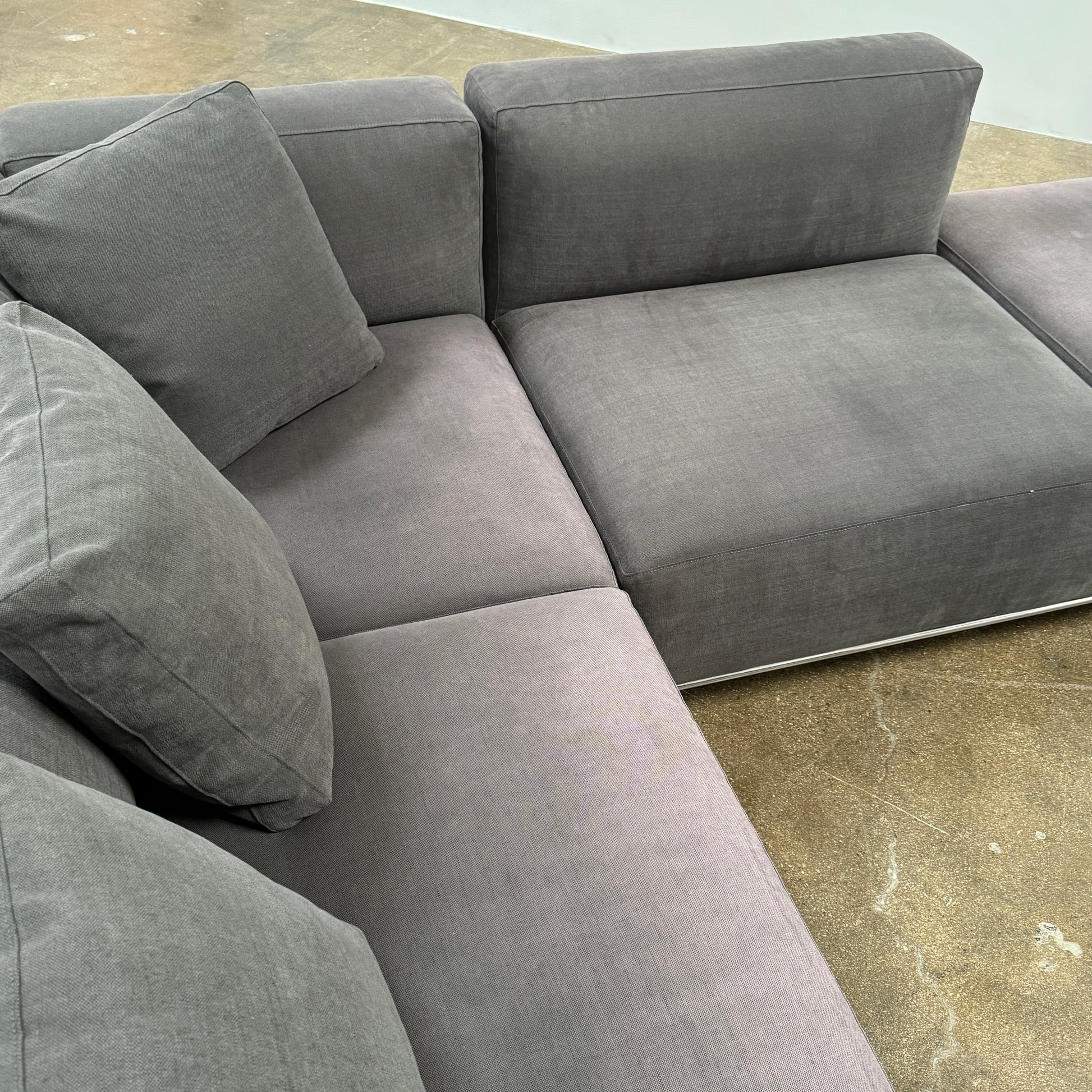The Cassina Mex Hi Modular Sofa by Cassina features dark gray recycled fiber upholstery, metal legs, multiple cushions, and an integrated wooden side table, shown against a white background.