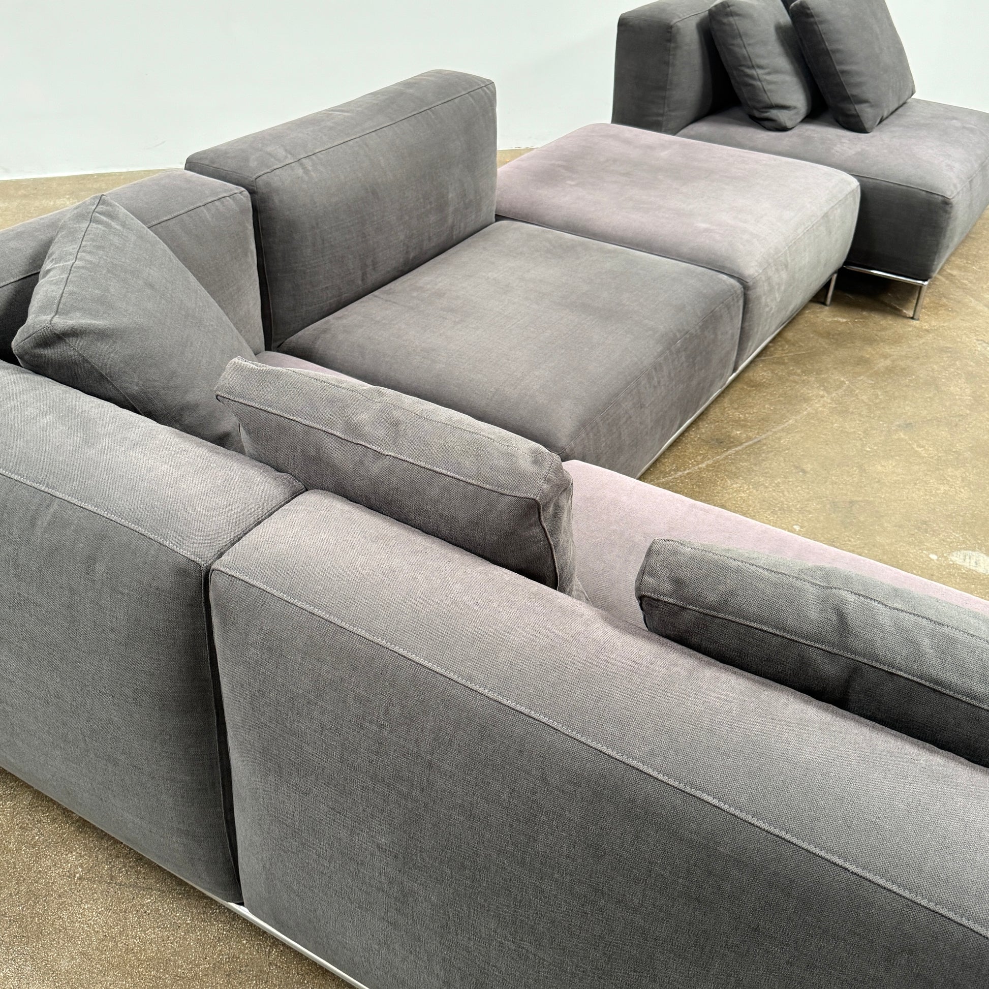 The Cassina Mex Hi Modular Sofa by Cassina features dark gray recycled fiber upholstery, metal legs, multiple cushions, and an integrated wooden side table, shown against a white background.