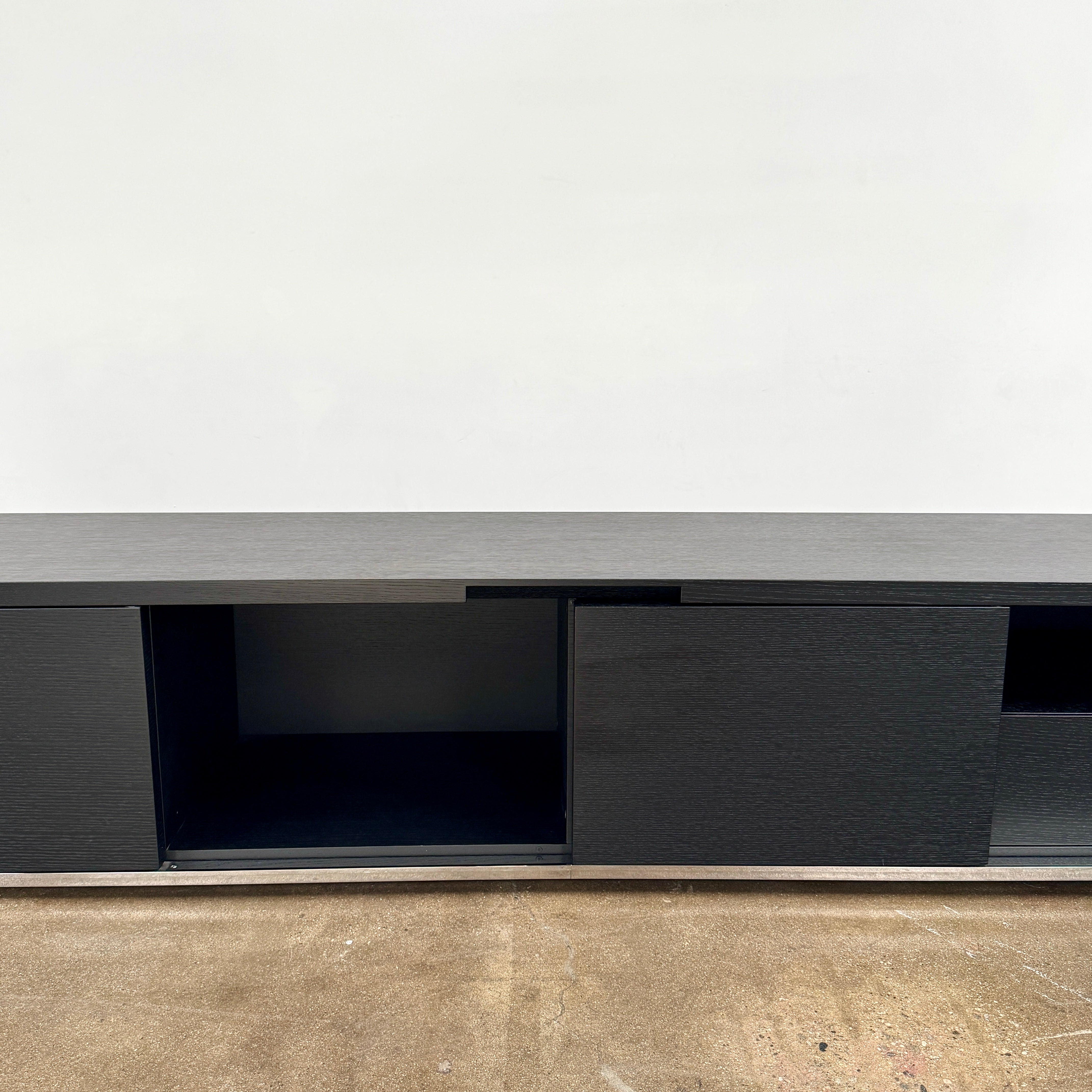 The Maxalto Mida Console by Maxalto is a modern black sideboard TV stand with three closed cabinets and two open shelves, shown on a concrete floor against a plain white background.