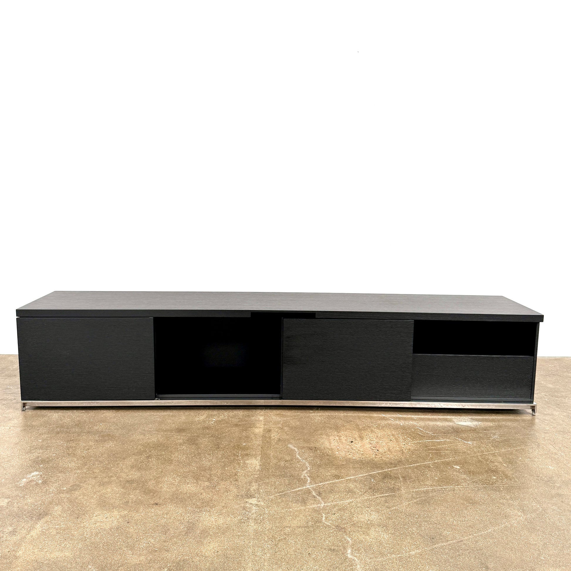 The Maxalto Mida Console by Maxalto is a modern black sideboard TV stand with three closed cabinets and two open shelves, shown on a concrete floor against a plain white background.