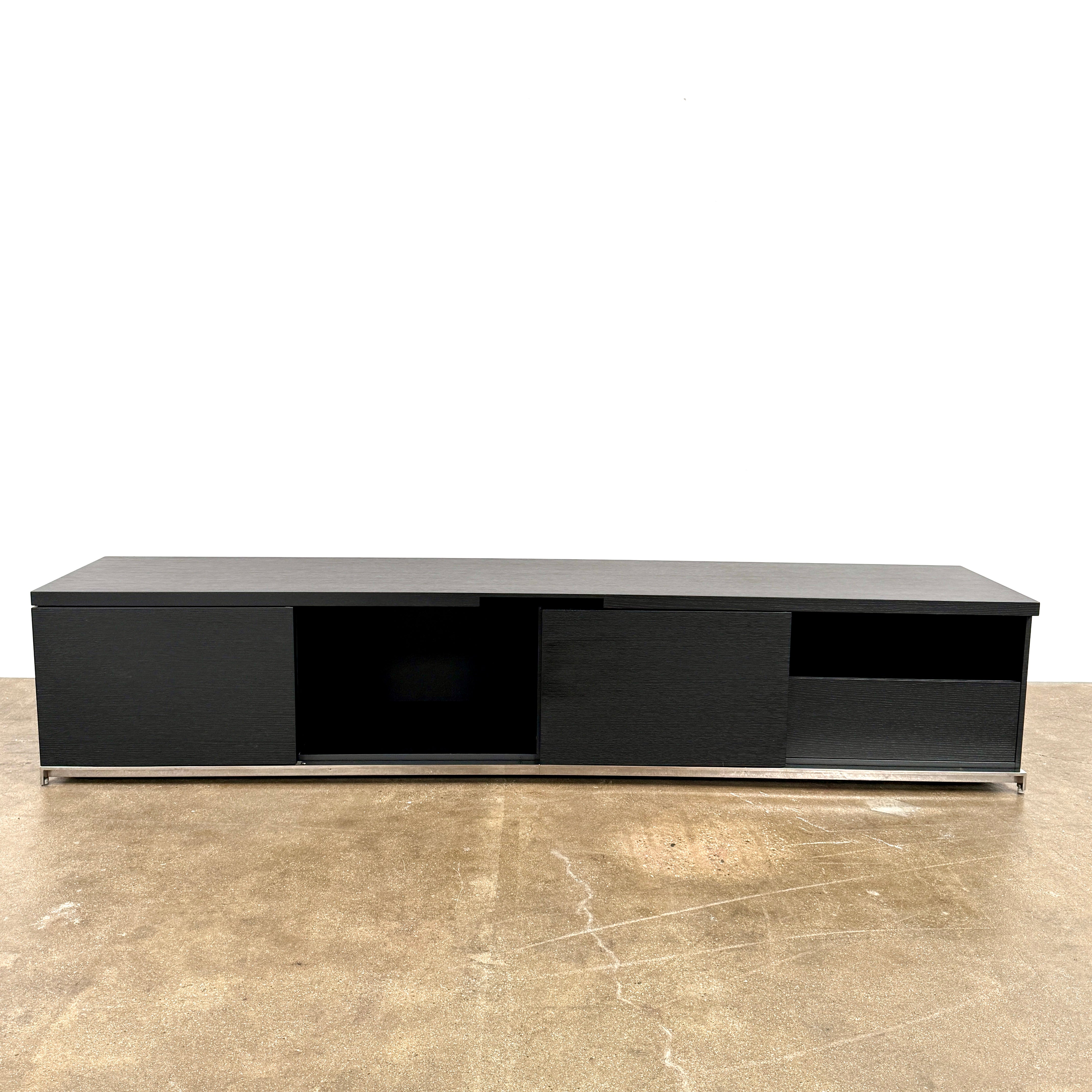 The Maxalto Mida Console by Maxalto is a modern black sideboard TV stand with three closed cabinets and two open shelves, shown on a concrete floor against a plain white background.