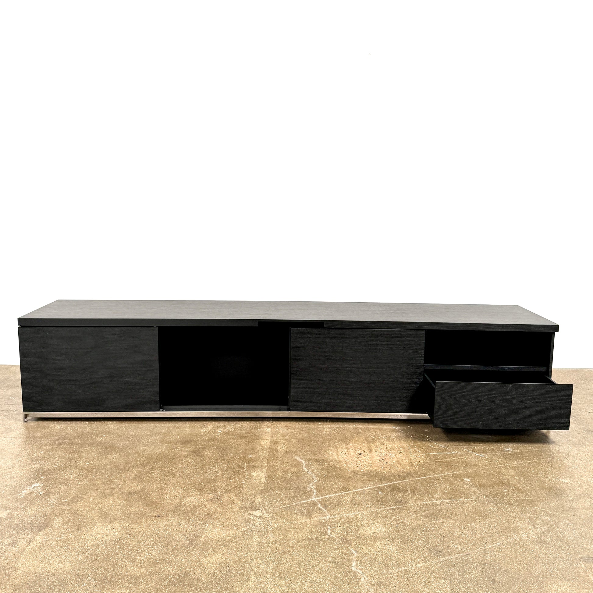 The Maxalto Mida Console by Maxalto is a modern black sideboard TV stand with three closed cabinets and two open shelves, shown on a concrete floor against a plain white background.