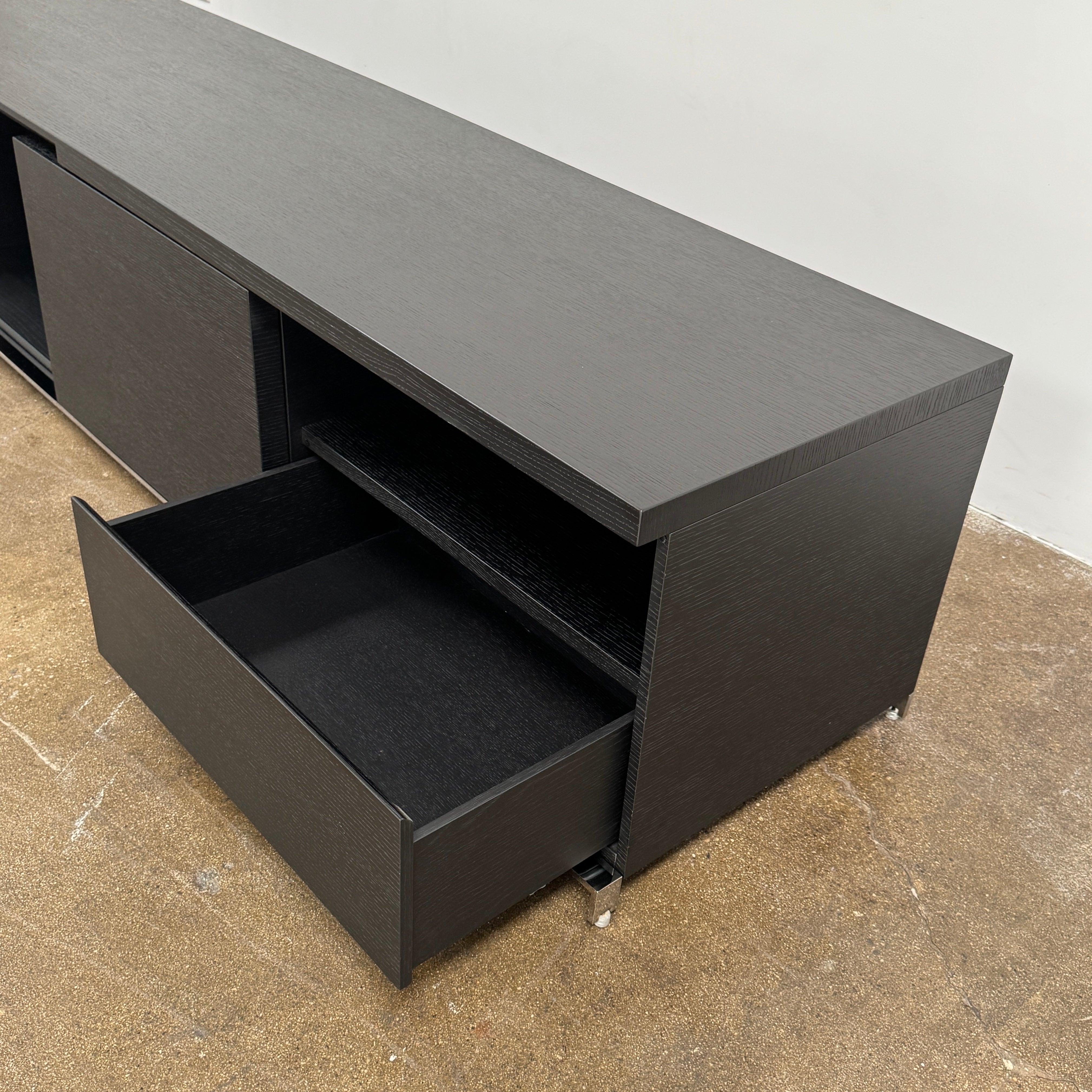 The Maxalto Mida Console by Maxalto is a modern black sideboard TV stand with three closed cabinets and two open shelves, shown on a concrete floor against a plain white background.